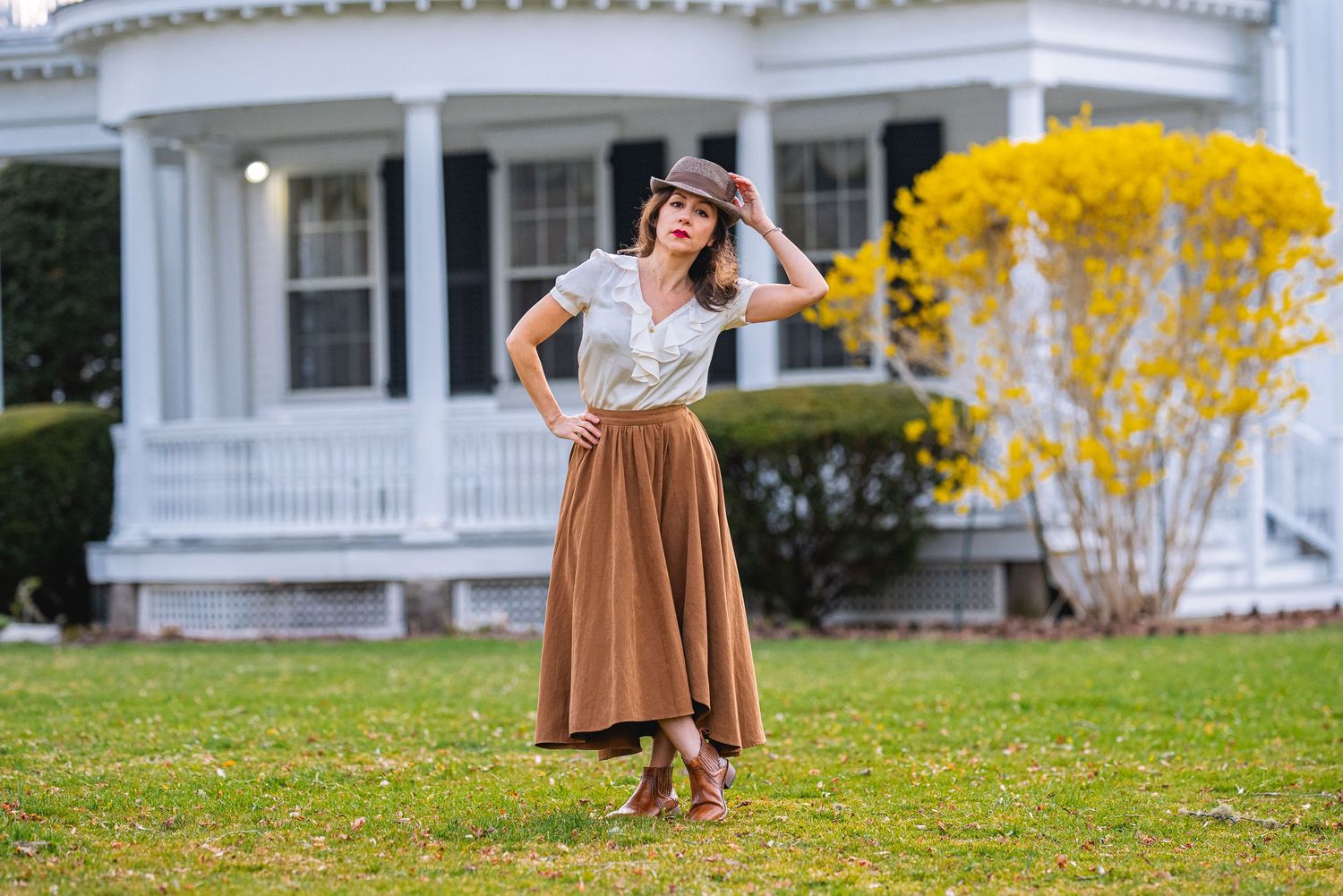 Woman in brown midi skirt and hat poses on lawn before white colonial house with yellow flowering shrubs.