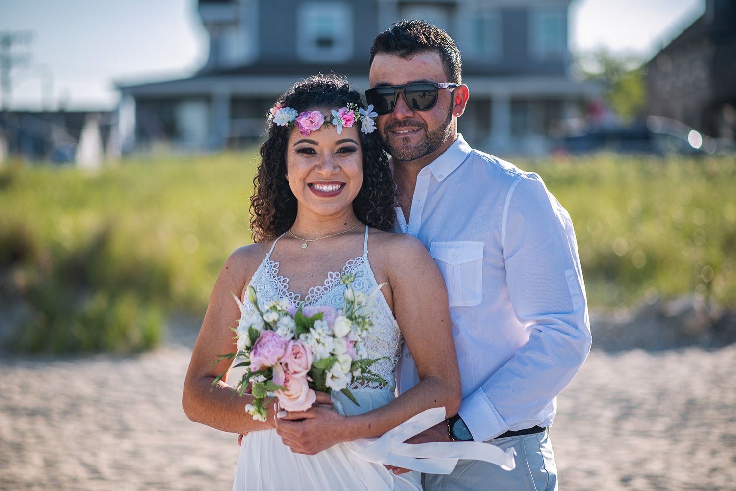 Smiling bride with pink flower crown holding bouquet embraced by groom in sunglasses on a sunny beach.