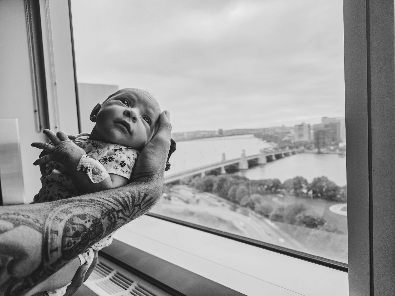 Tattooed arm cradling a sleeping newborn baby beside a large window overlooking a river, bridge and city skyline in black and white.