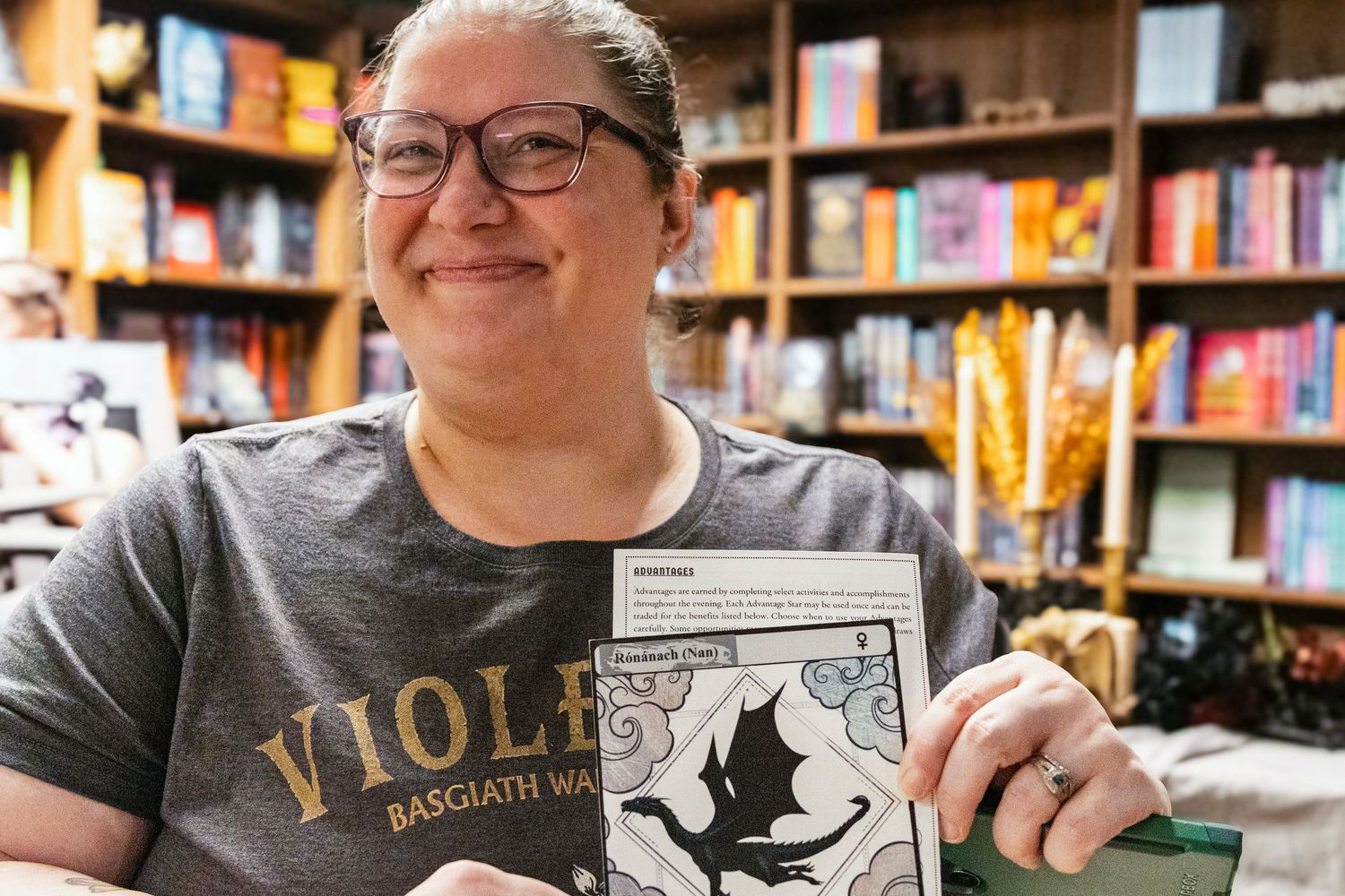 Woman in Viola t-shirt smiling and holding a card in a cozy bookstore with colorful books on shelves.
