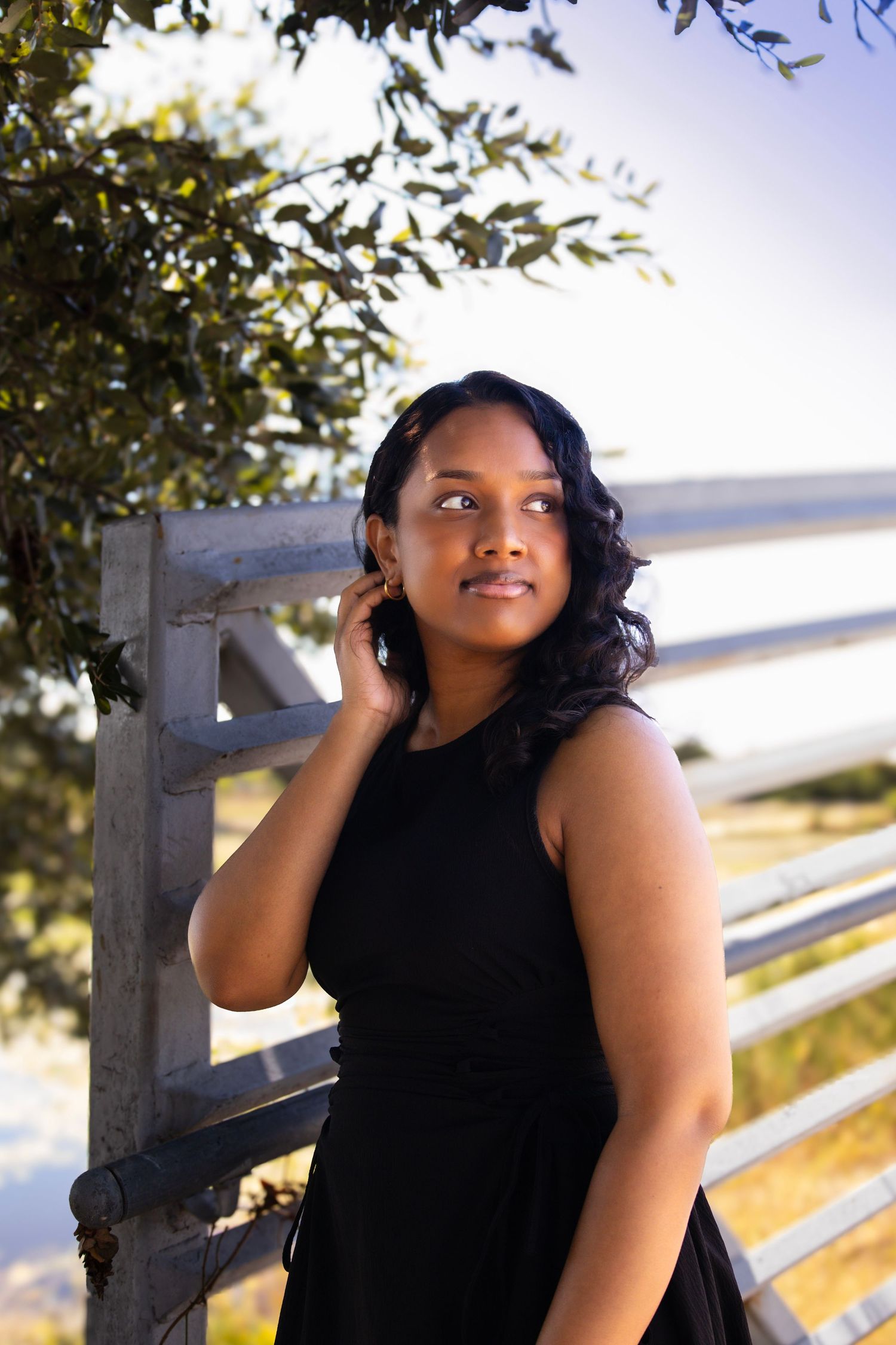 Young Black woman in black dress stands by wooden fence near water, gazing upward with hand touching hair.