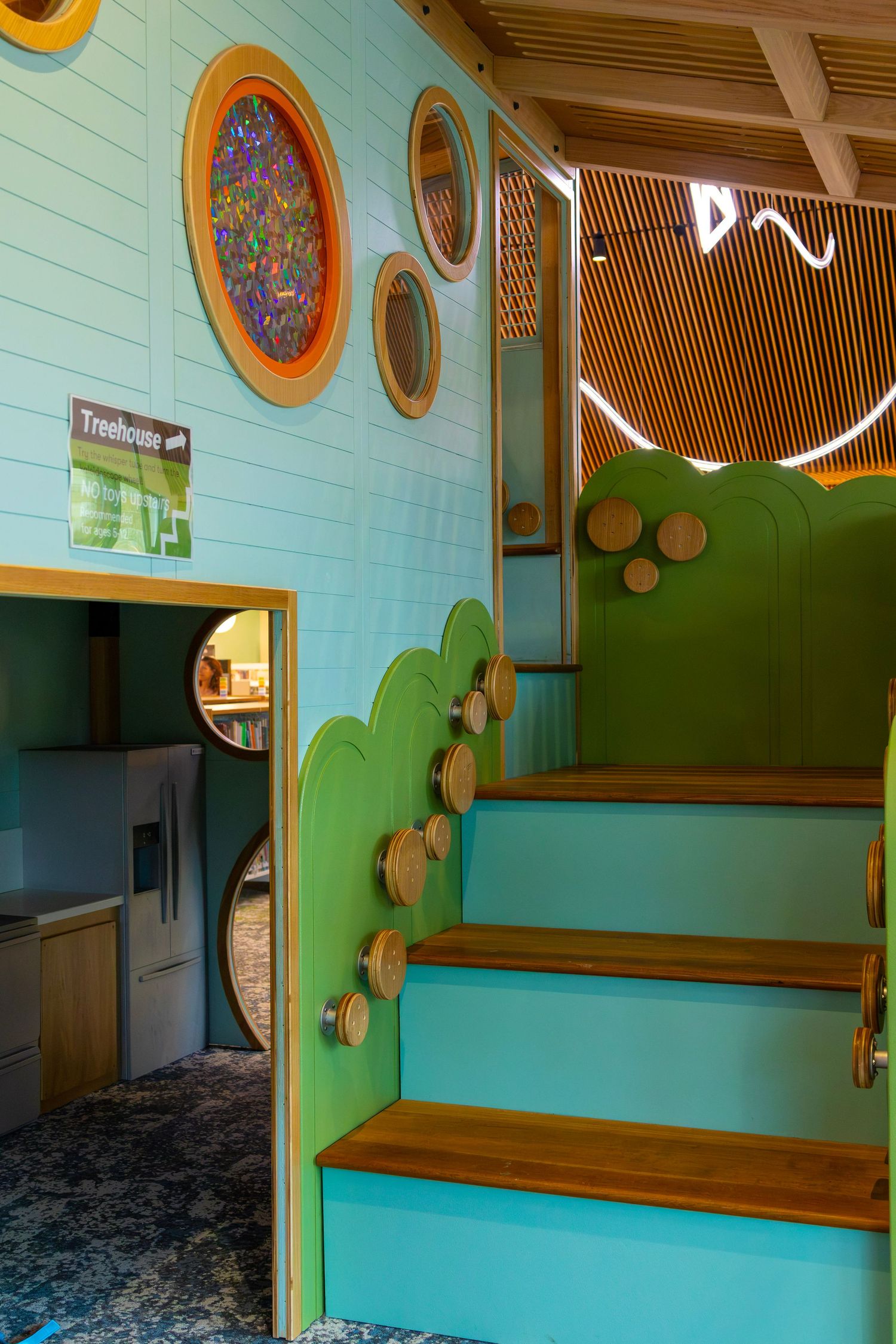 Whimsical treehouse-themed library play area with green stairs, circular windows, and warm wood accents.