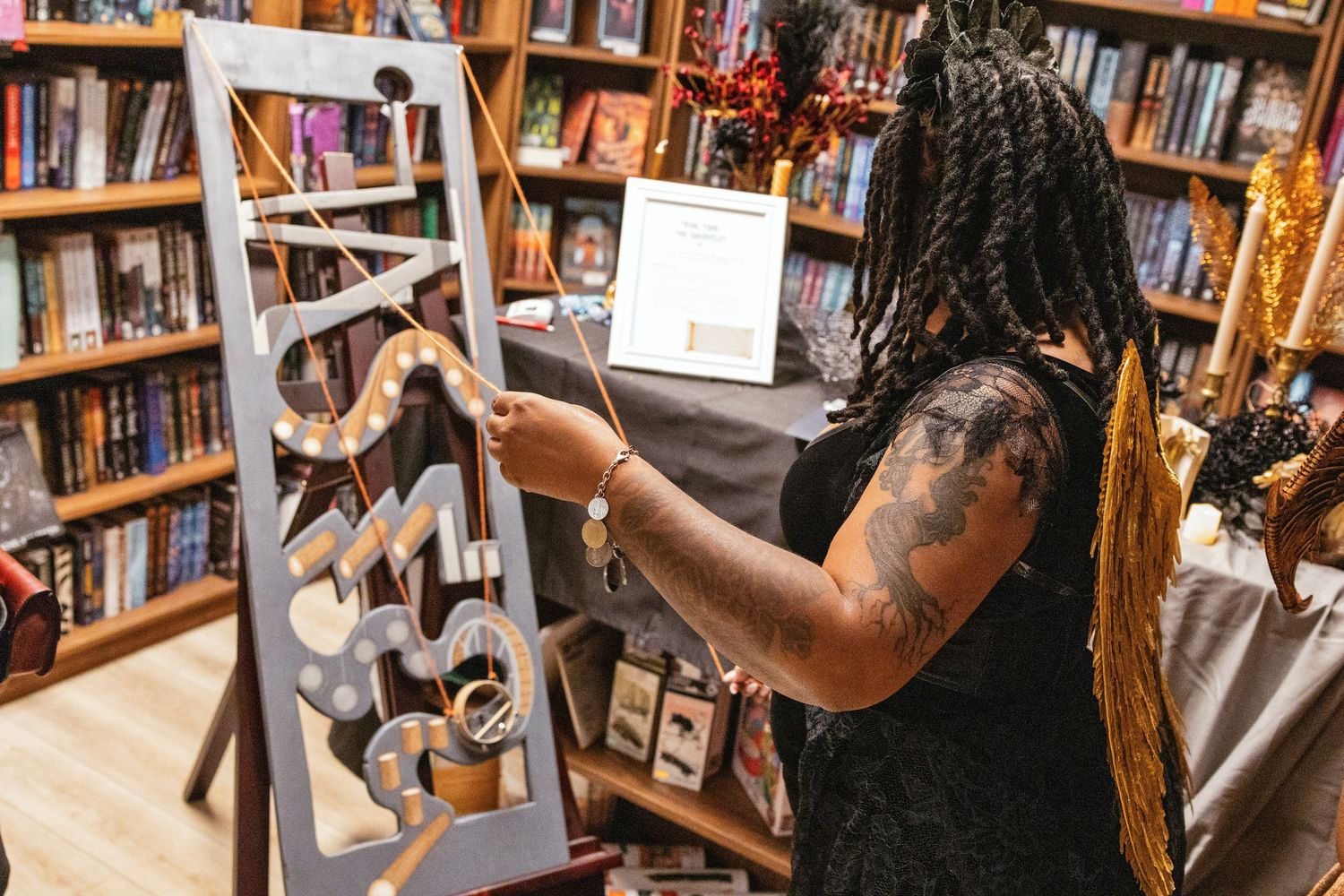 Tattooed person examines wooden snake-shaped jewelry display in a bookstore filled with shelves.