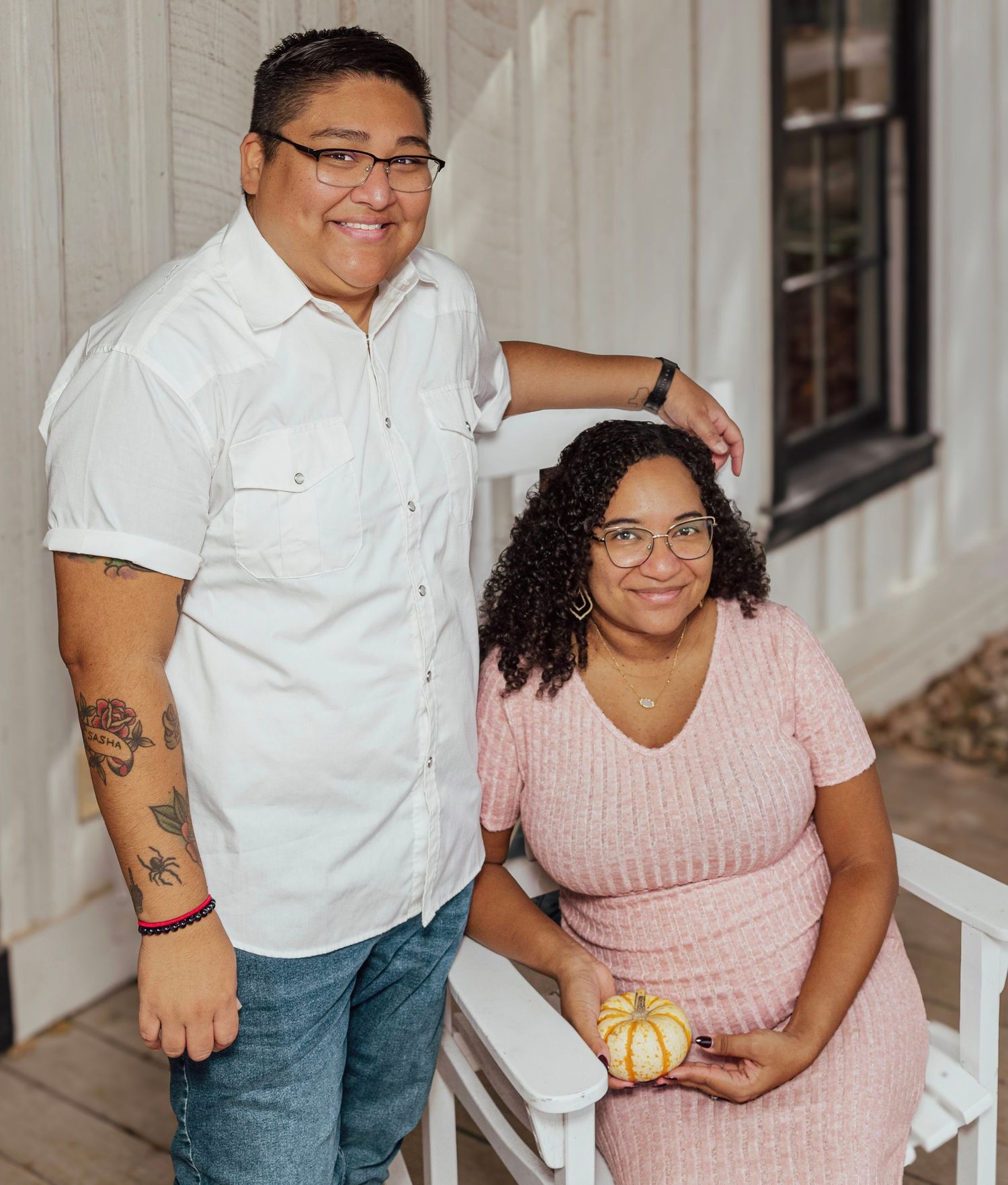 Two people smiling on a porch, one standing in a white shirt with tattoos, one seated in pink holding a small pumpkin.