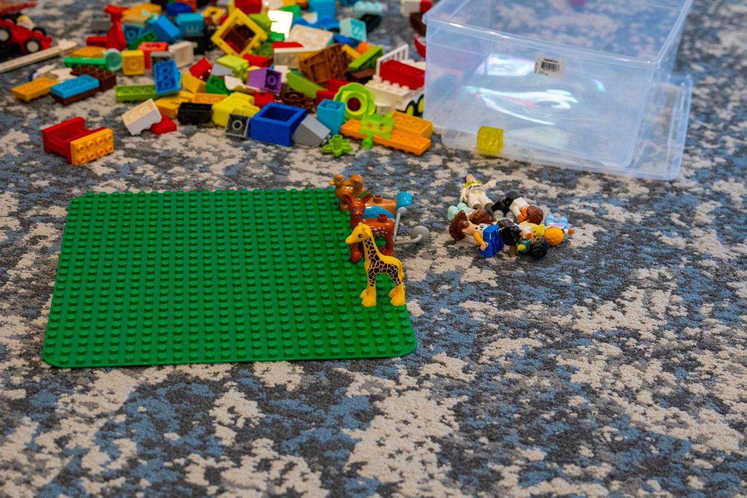 Colorful LEGO Duplo bricks scattered near a green baseplate with small figures on a gray carpet.