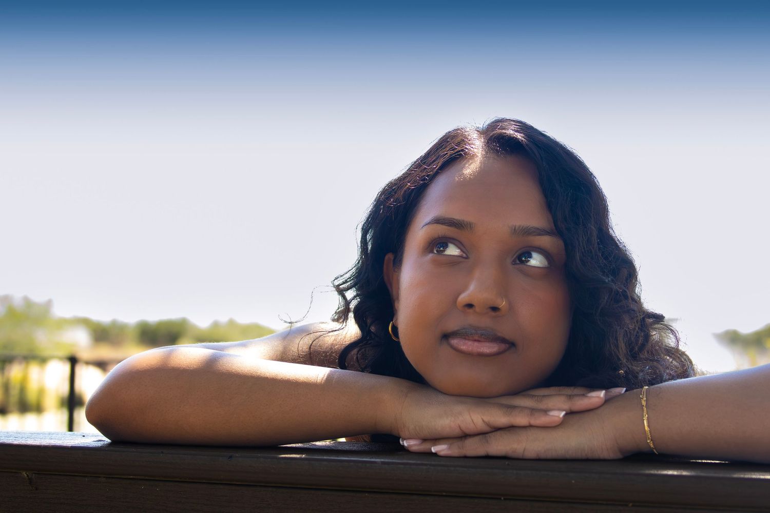 Young Black woman resting her chin on folded hands, gazing thoughtfully upward outdoors on a sunny day.