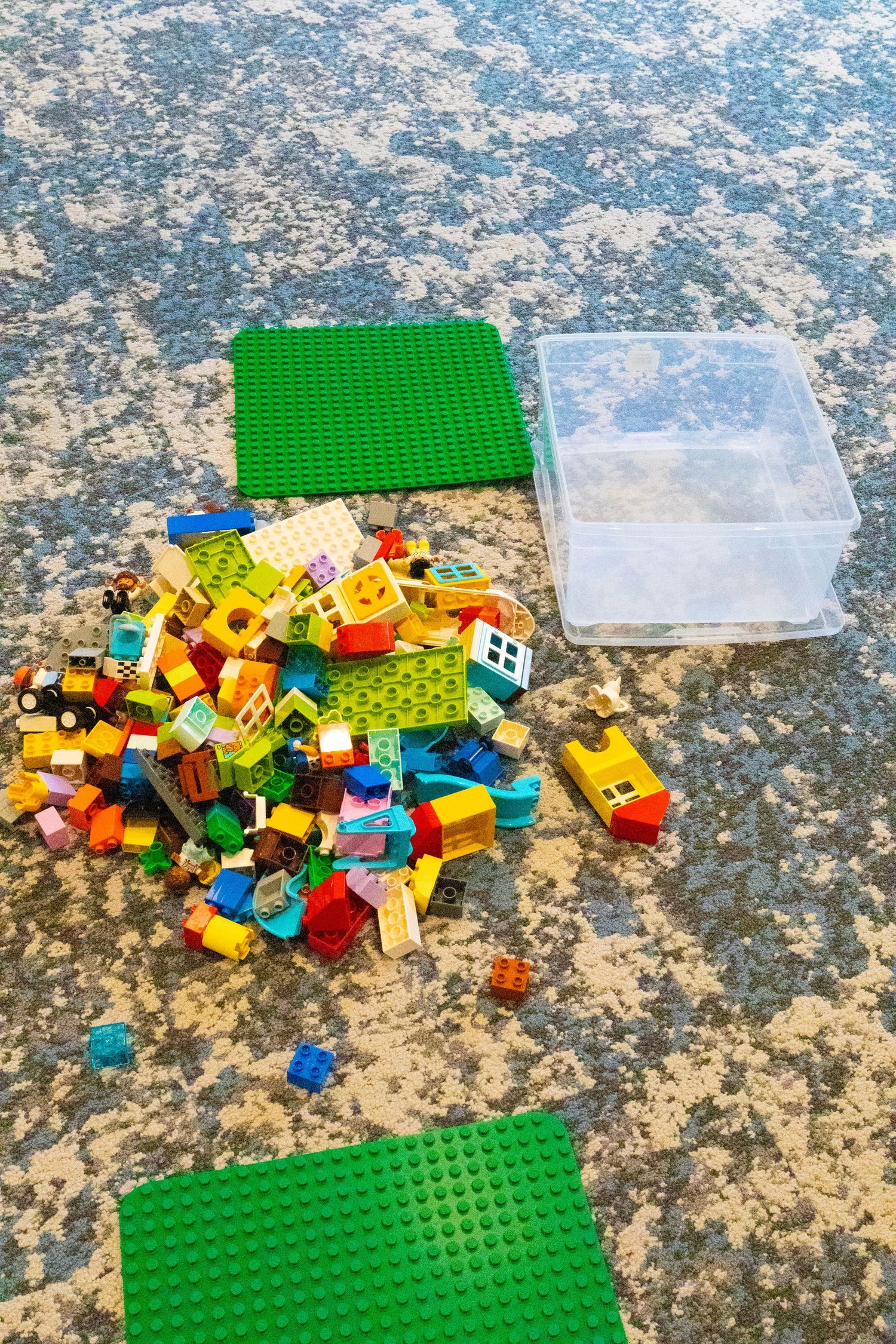 Scattered colorful LEGO Duplo building blocks with green base plates and clear storage container on granite surface.