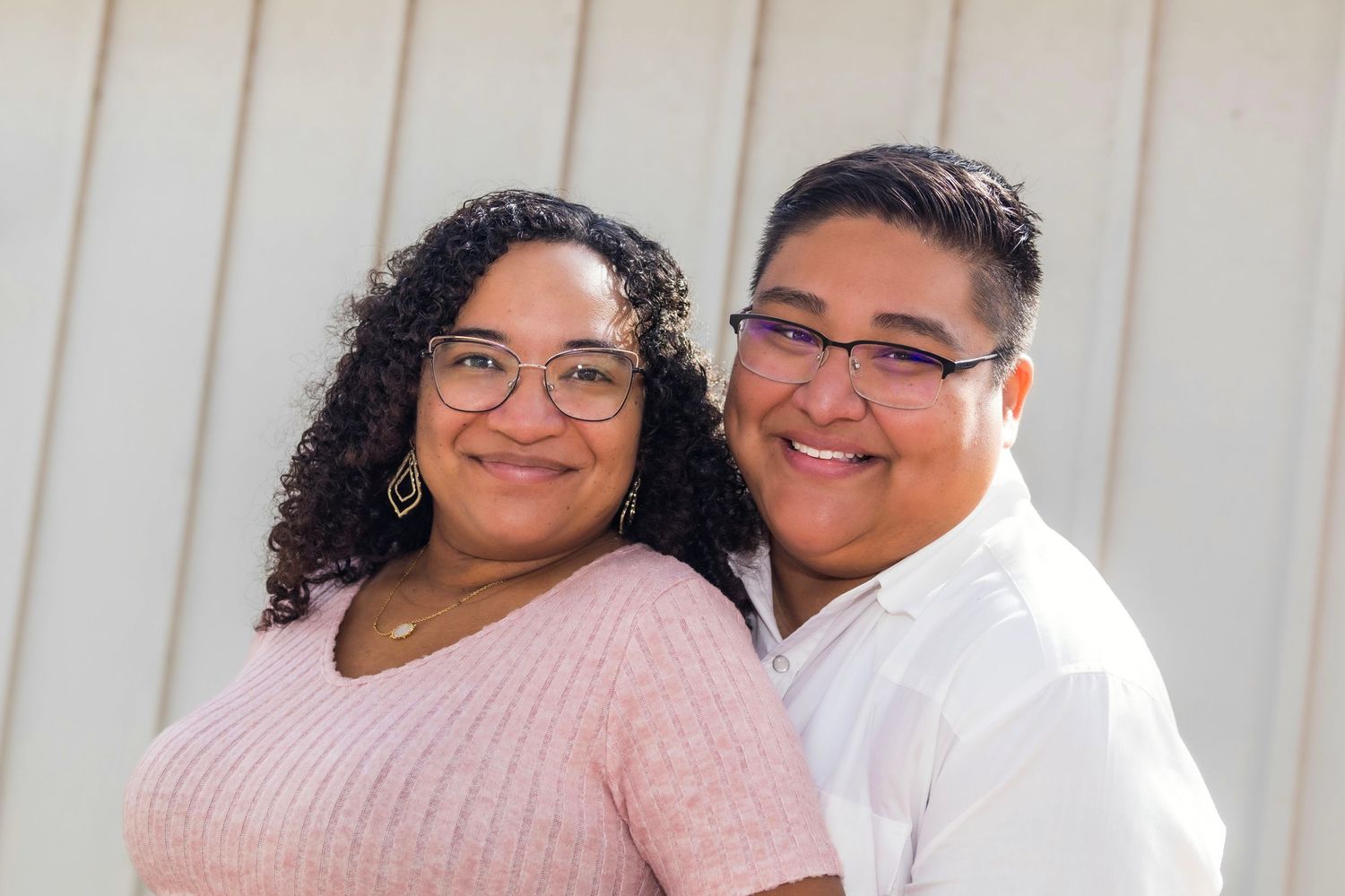 Two women wearing glasses smile together in a close portrait, one in pink and one in white, outdoors.
