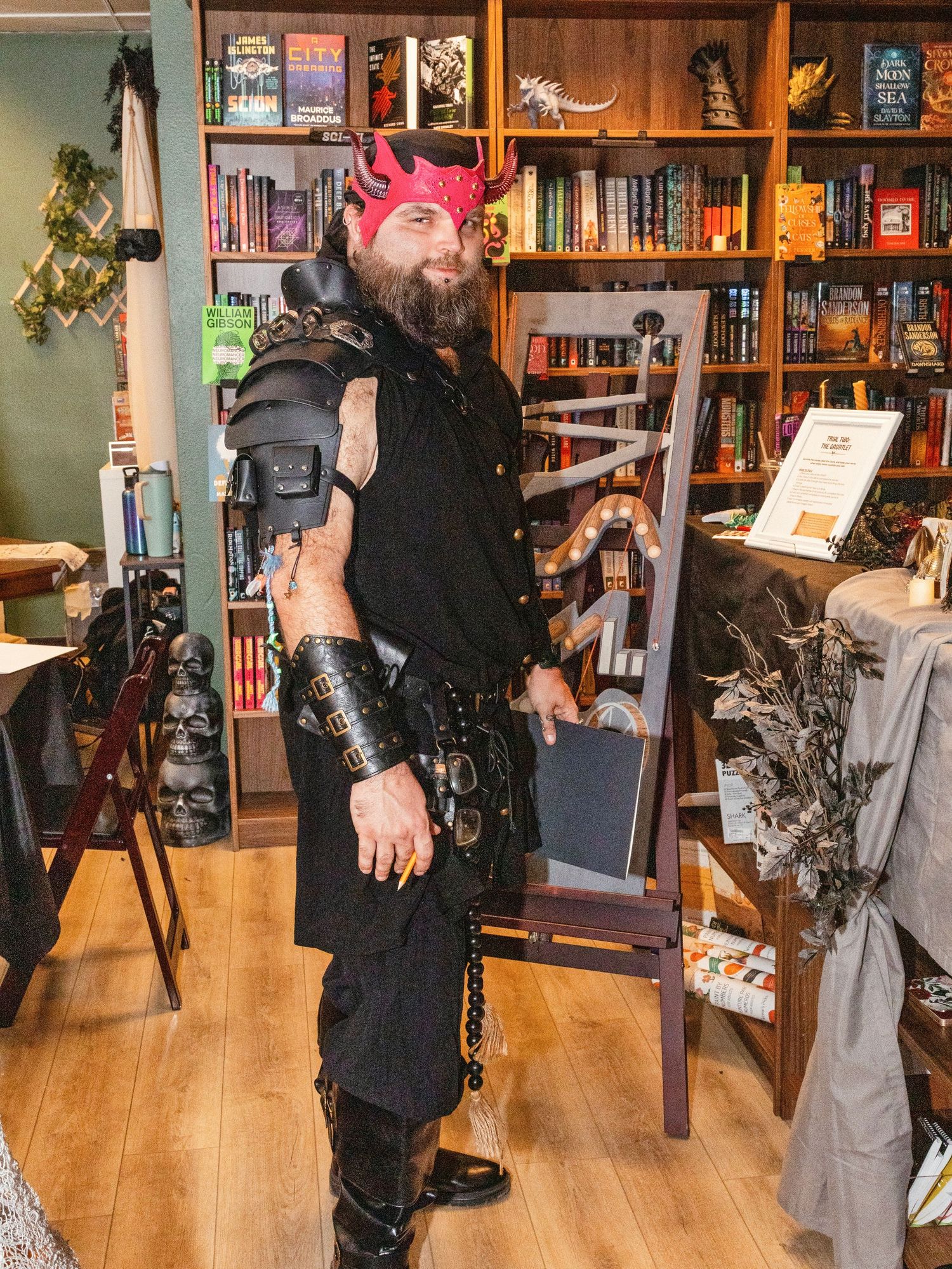Bearded man in black medieval armor with red devil horns poses in a fantasy-themed bookstore.