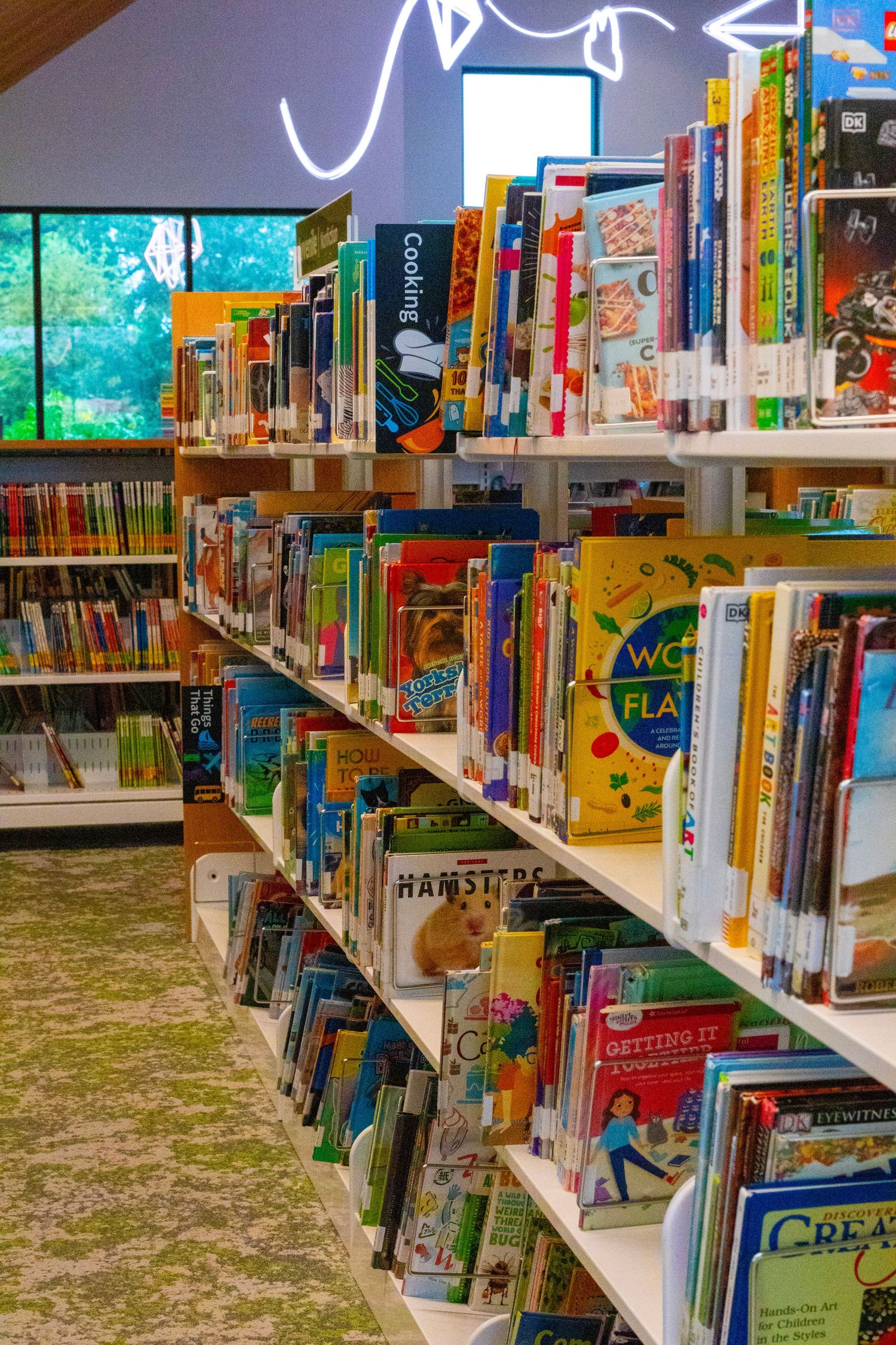 Bright childrens library shelves filled with colorful books including cooking and activity titles near large windows.