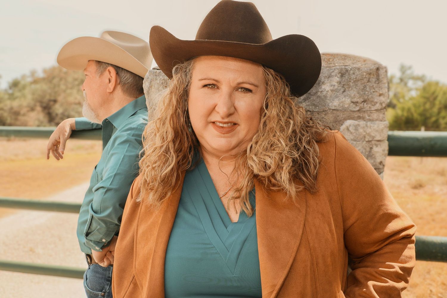 Woman in brown cowboy hat and teal top with orange jacket smiles outdoors near a fence.