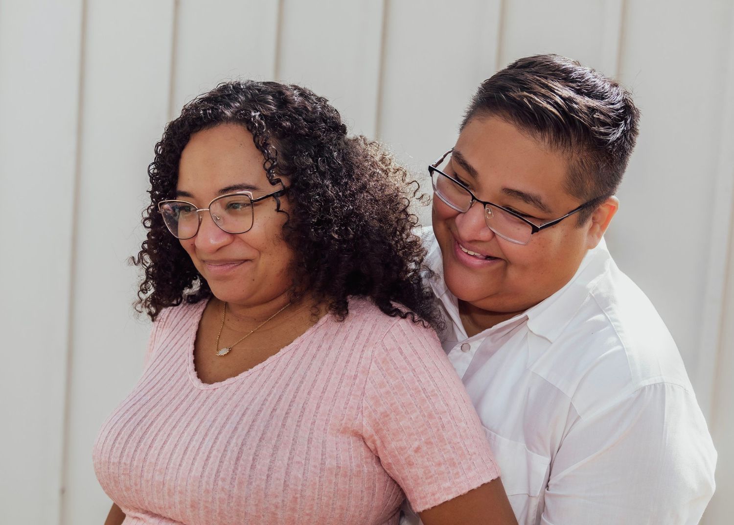 Two people wearing glasses smile warmly together outdoors, one in a pink knit top and the other in a white shirt.