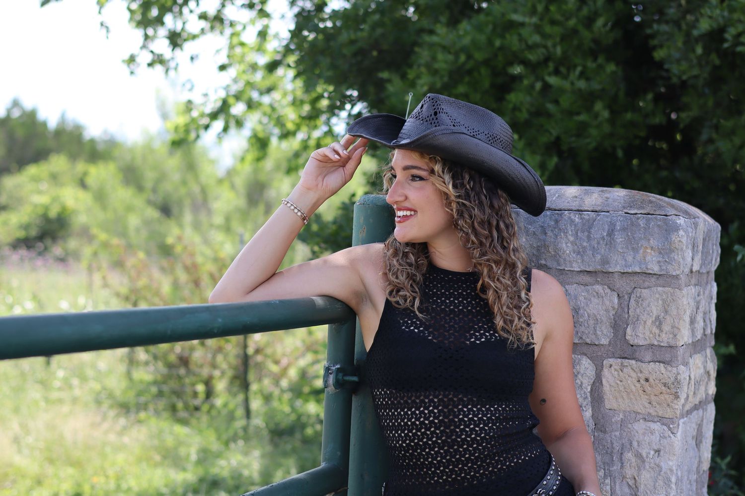 Photo by Lexy Hiam Woman in a black cowboy hat and black dress smiling while leaning on a green fence outdoors.