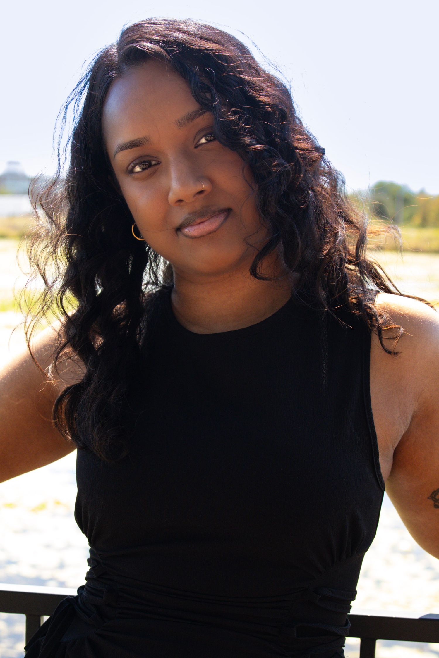 A young Black woman with long curly hair wearing a black sleeveless top poses outdoors near a waterfront.