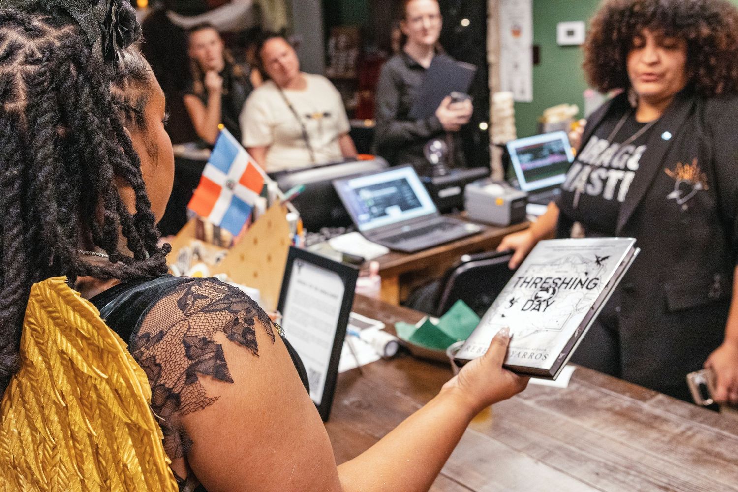 Woman holds an open book at a lively bookstore event with people gathered in the background.
