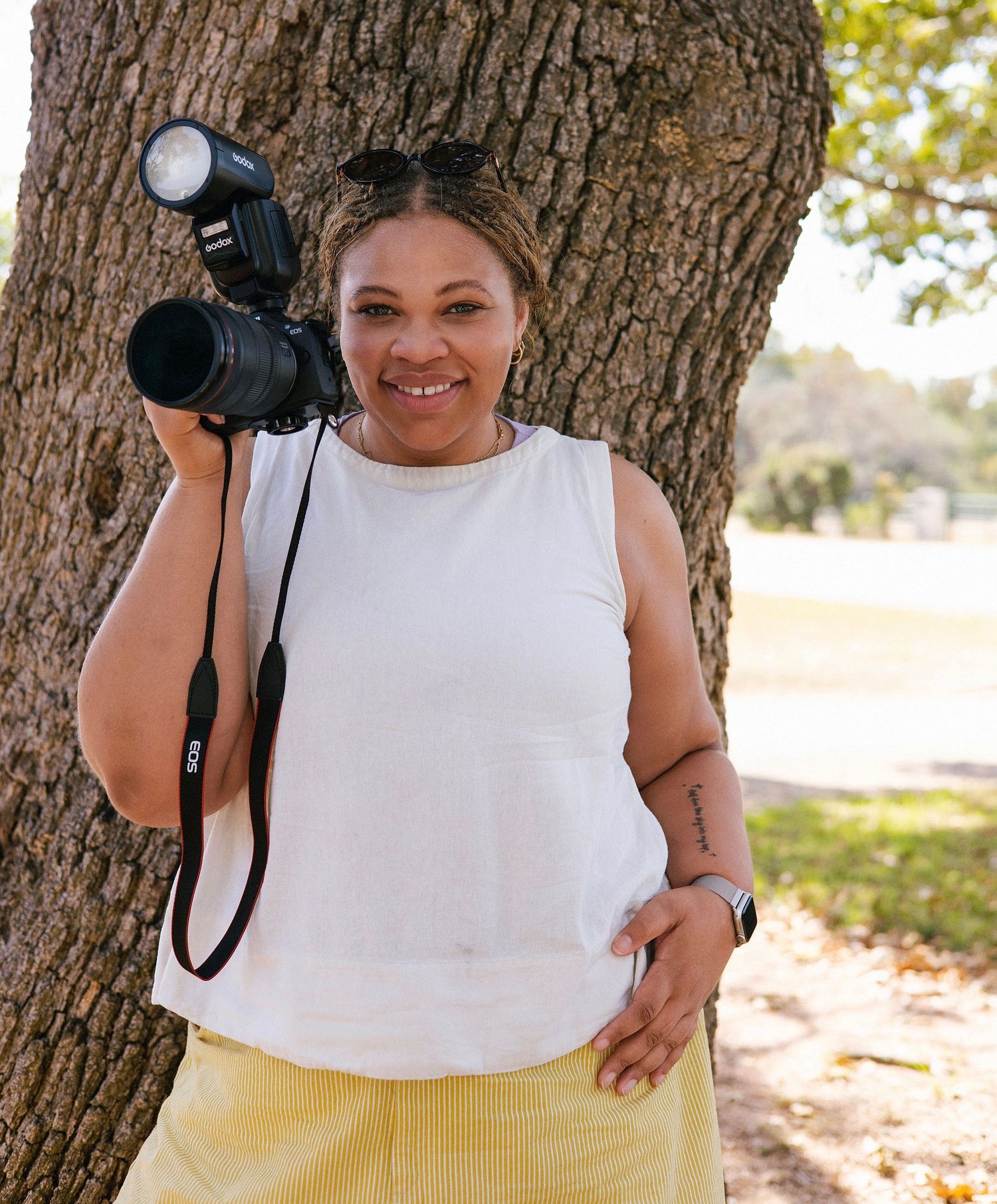 A smiling woman holds a DSLR camera with flash outdoors, leaning against a tree in a white sleeveless top and yellow pants.