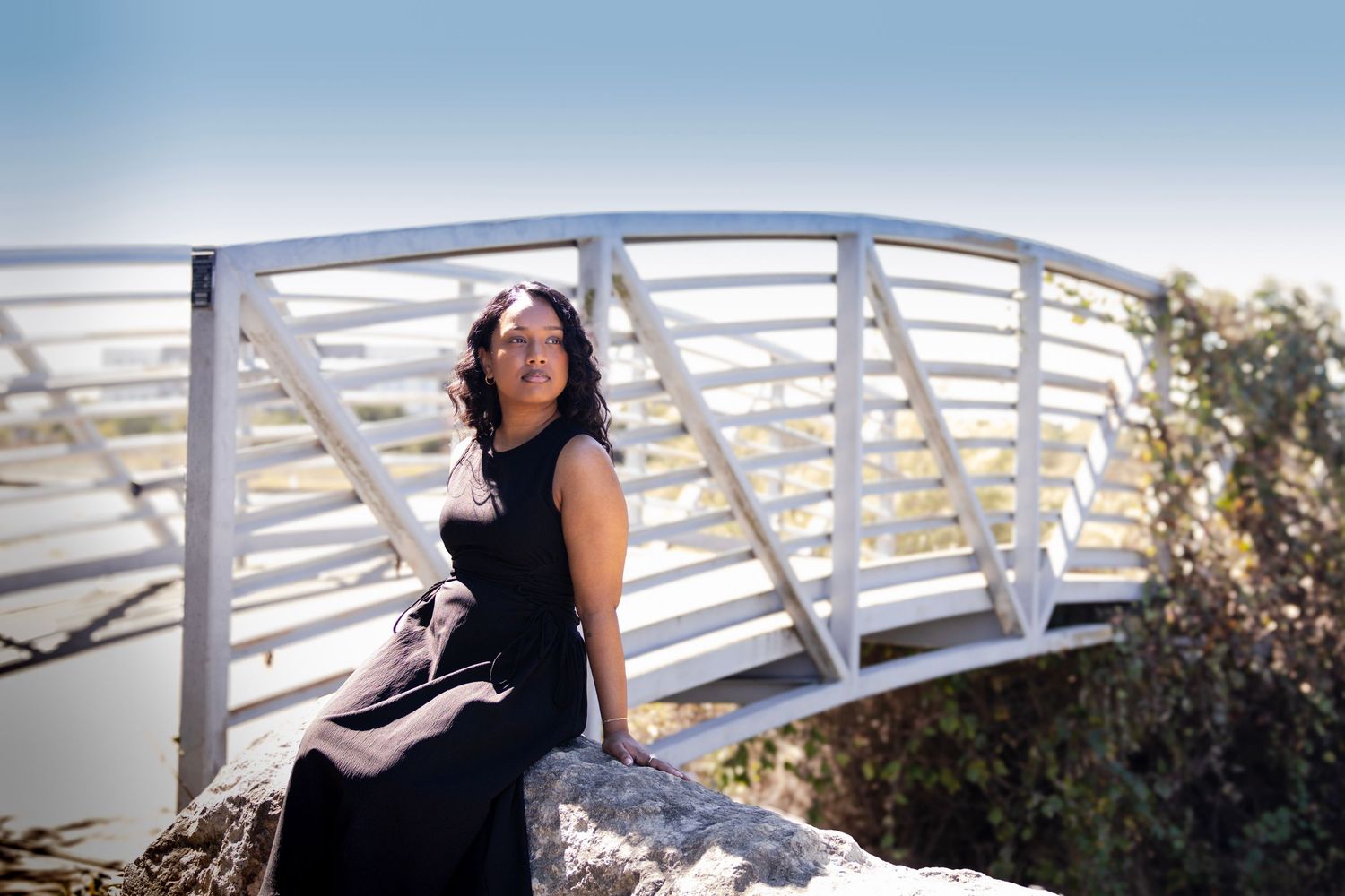 Woman in black dress sitting on rocks by a white arched bridge outdoors on a sunny day.