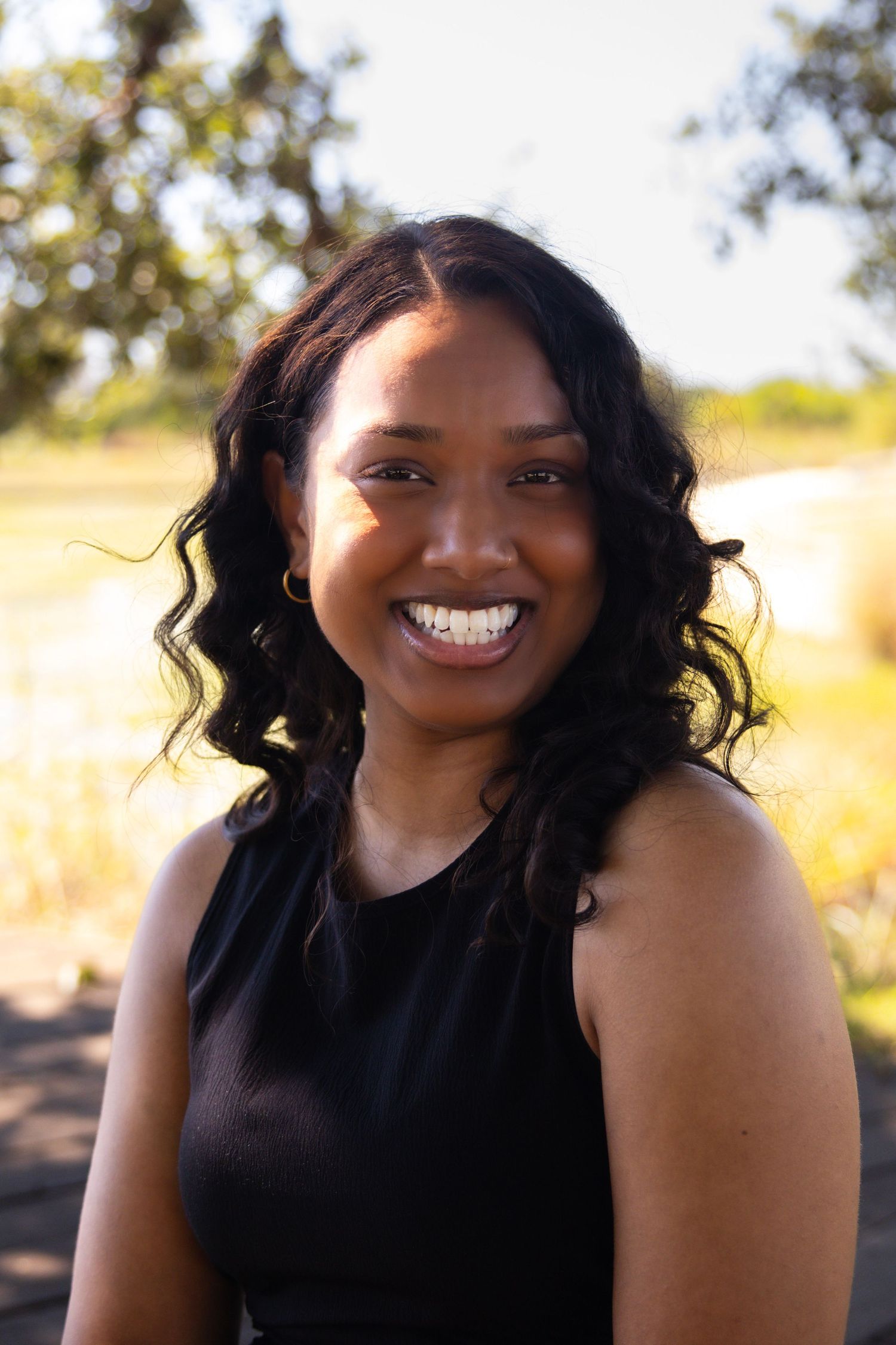 Smiling young Black woman with curly hair wearing a black sleeveless top outdoors on a sunny day.
