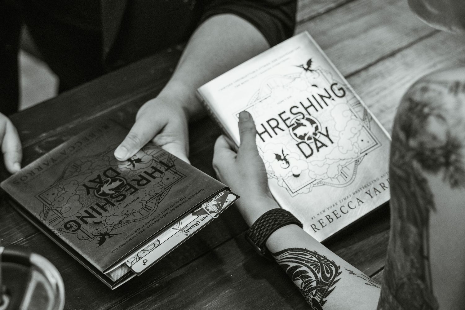 Two people examining copies of Threshing Day book by Rebecca Ray on a wooden table in black and white.