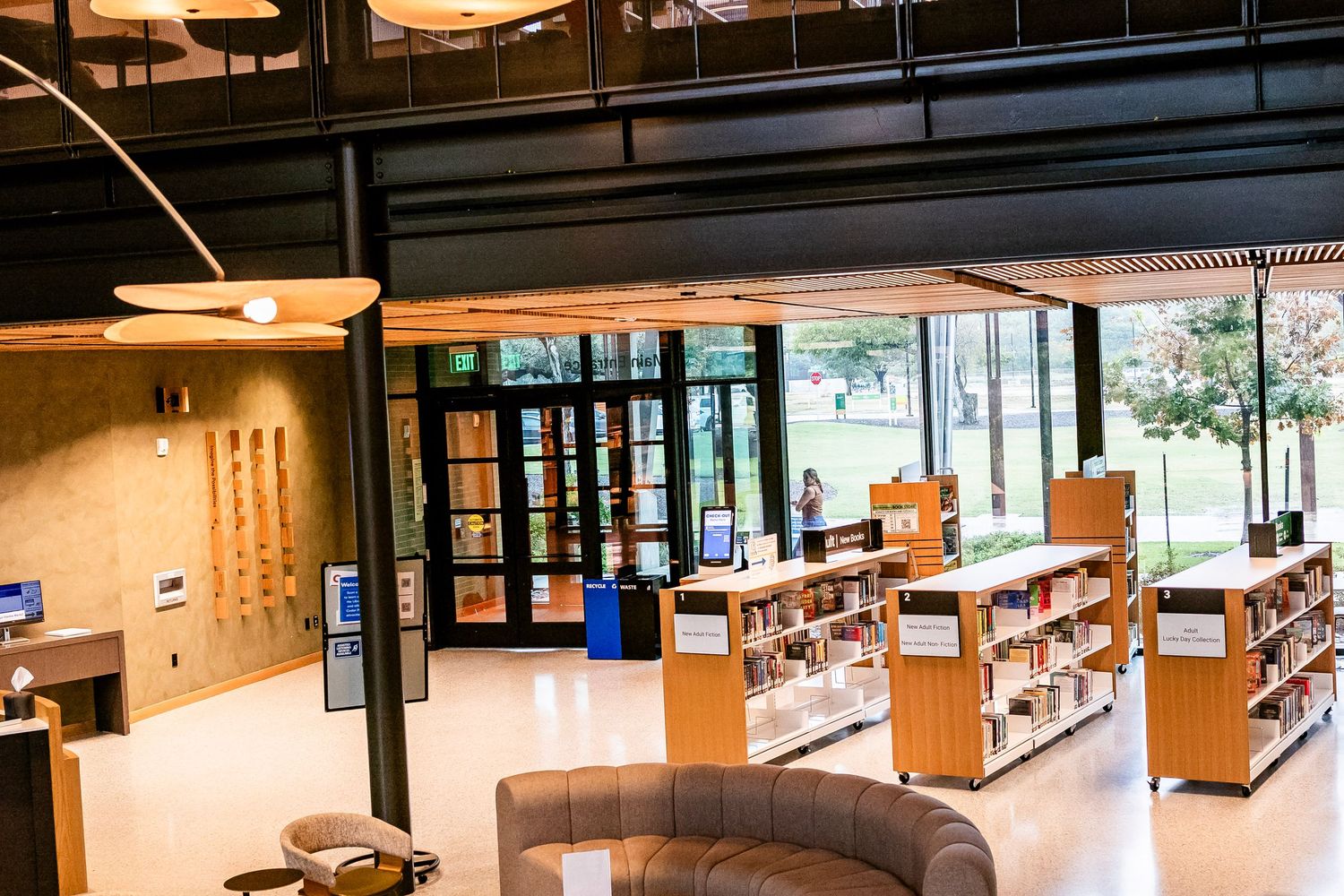 Modern library interior with wooden bookshelves, curved sofa seating, and large glass entrance doors overlooking a green courtyard.