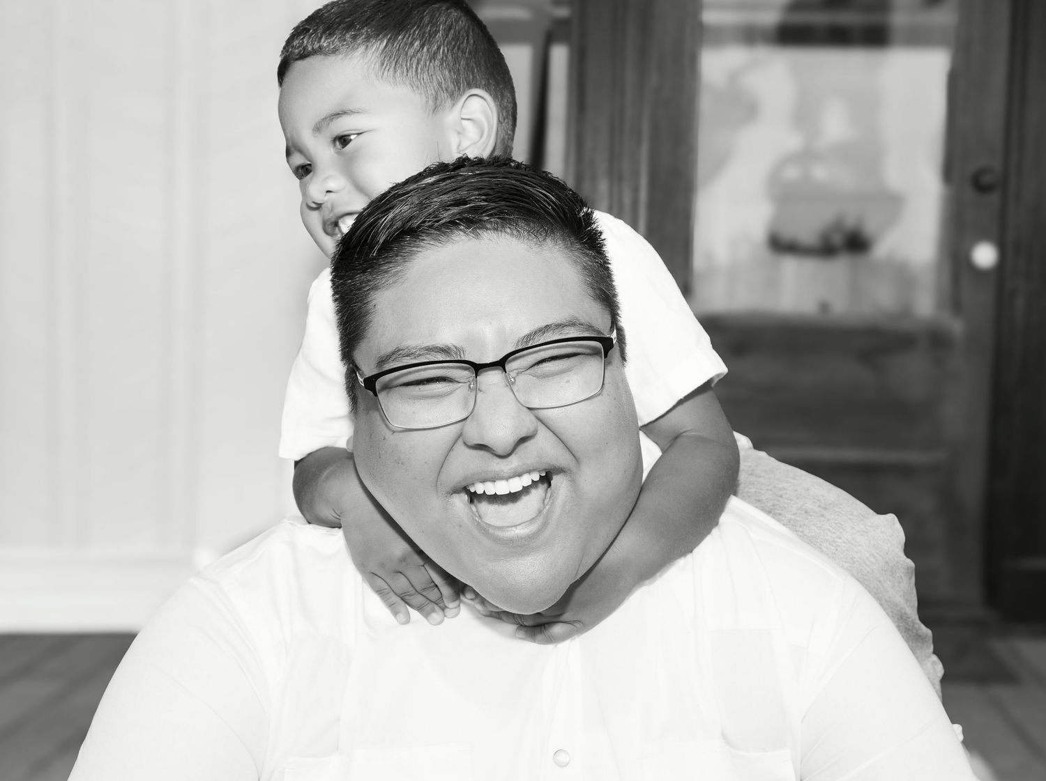 Black and white photo of a laughing man wearing glasses with a young boy sitting on his shoulders smiling.