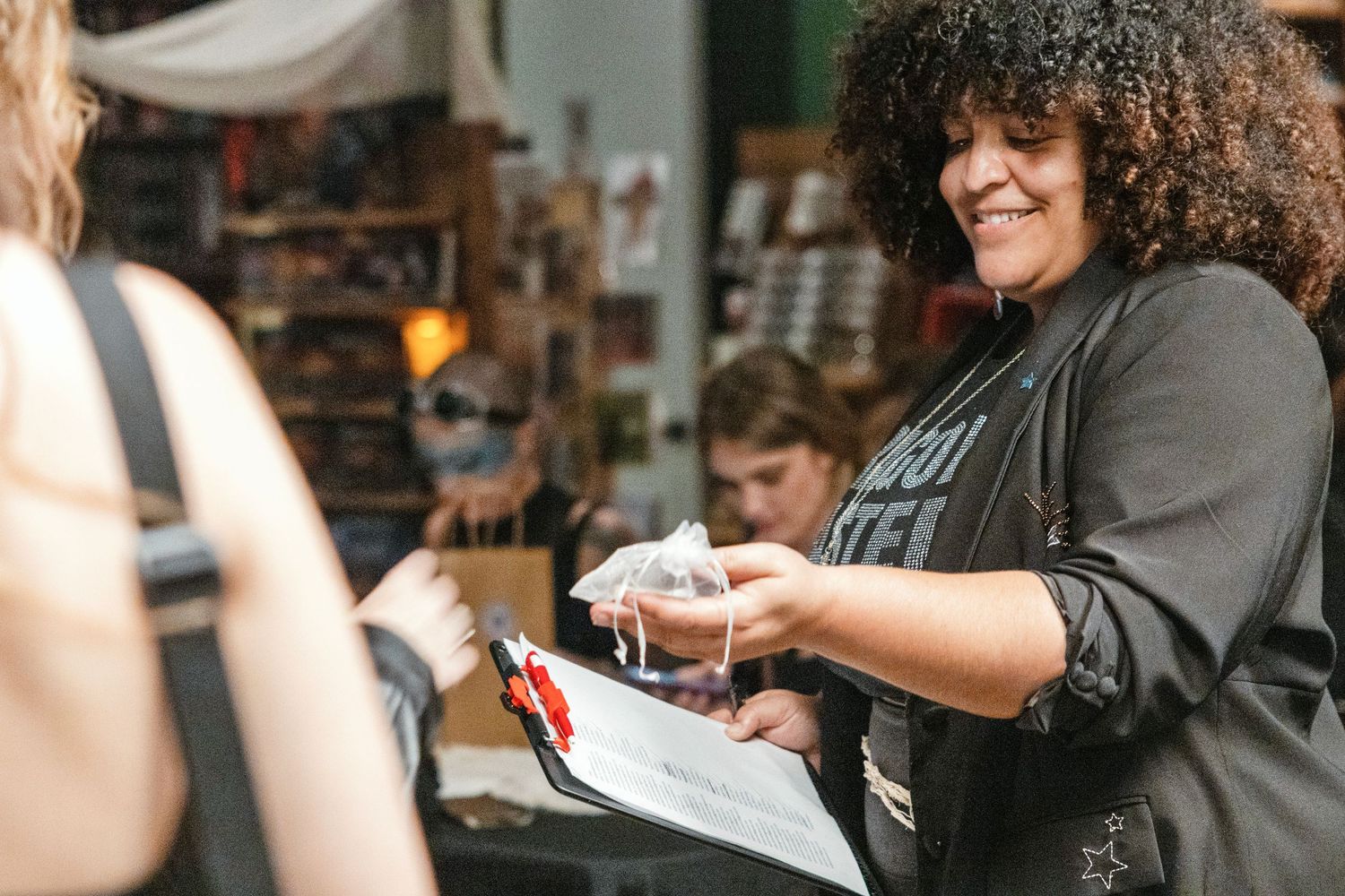 Smiling curly-haired woman hands a packaged item to a customer at an indoor market event.