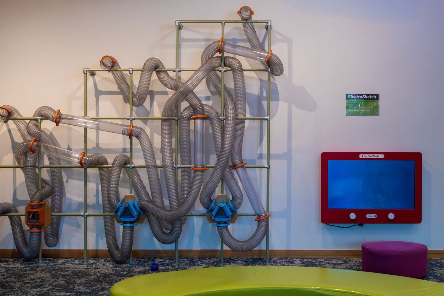 Interactive tube wall sculpture with maze-like pipes beside a red-framed blue screen in a childrens library play area.