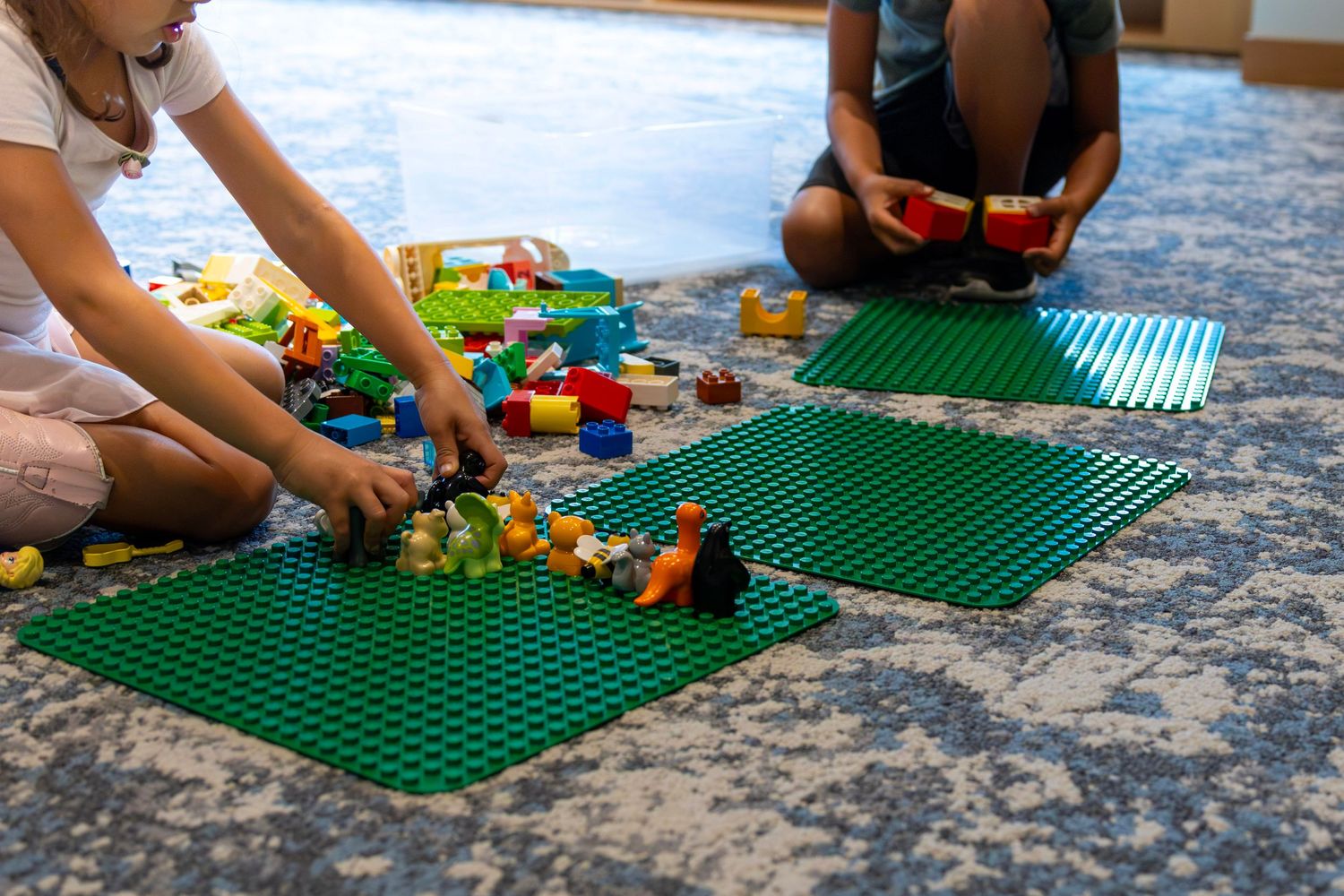 Children playing with colorful LEGO Duplo blocks on green baseplates on a carpet floor.