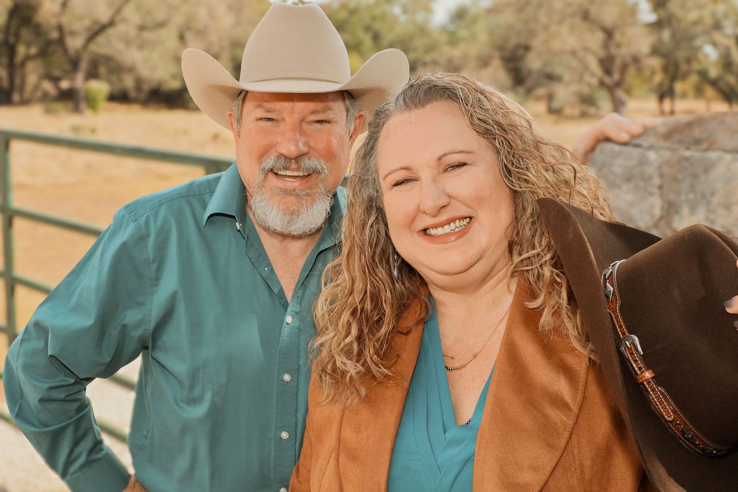 Smiling couple outdoors on a ranch, man in cowboy hat and teal shirt, woman in brown jacket.
