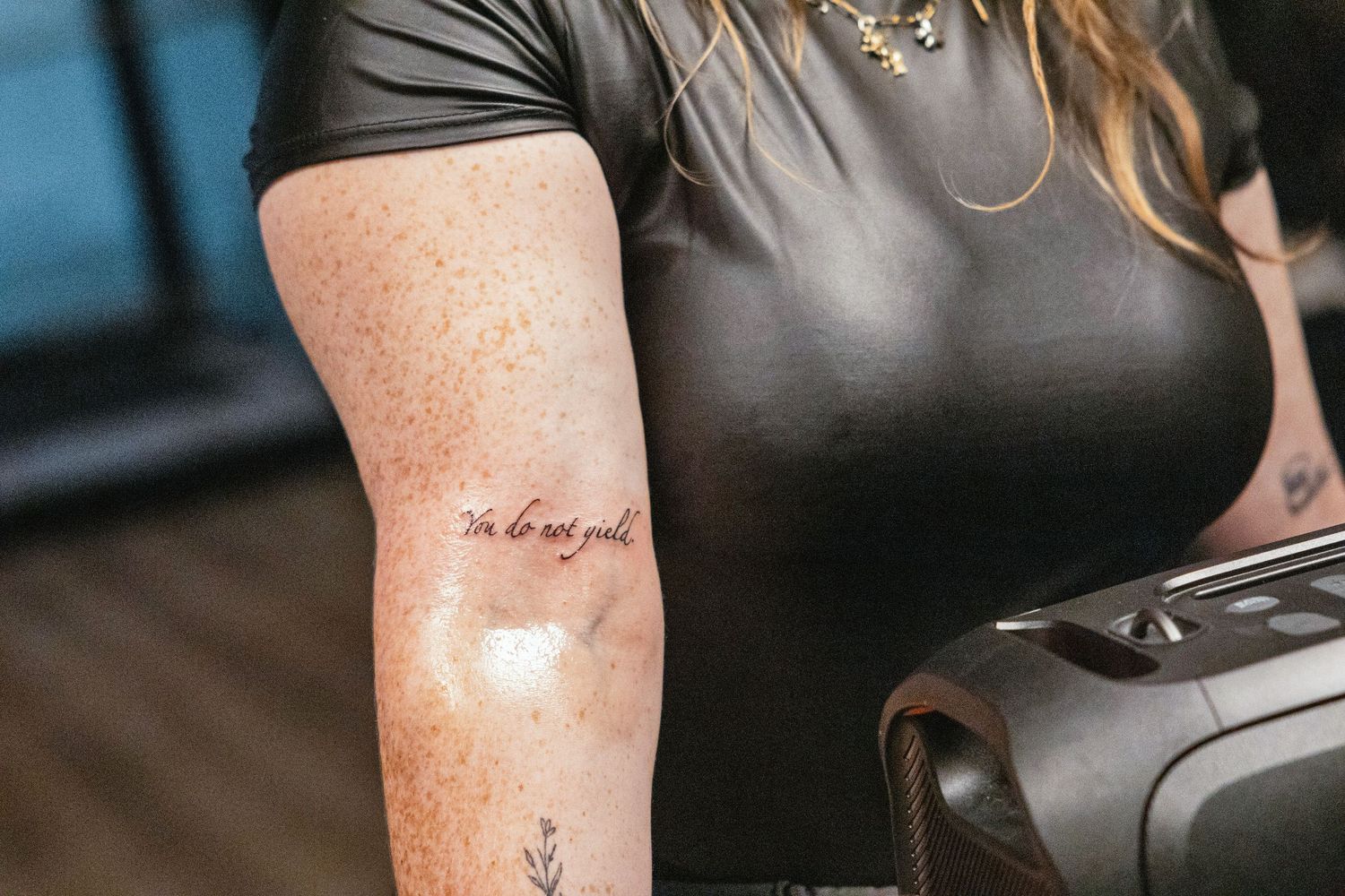 Close-up of a woman's arm with a cursive script tattoo and small floral tattoo, wearing a black top.