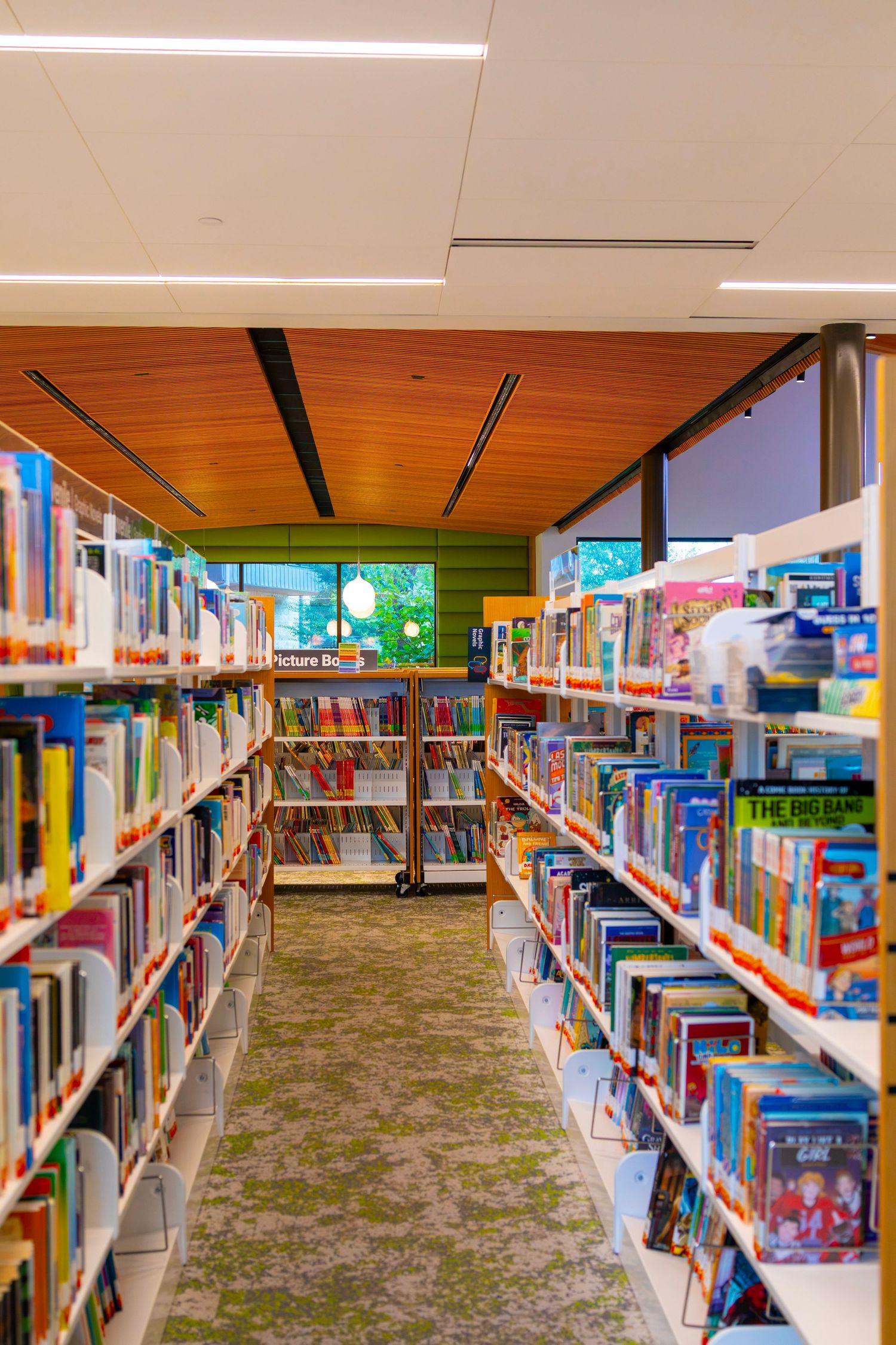 Colorful children's library aisle with shelves full of books, green carpet, wood ceiling, and a bright screen visible at the end.