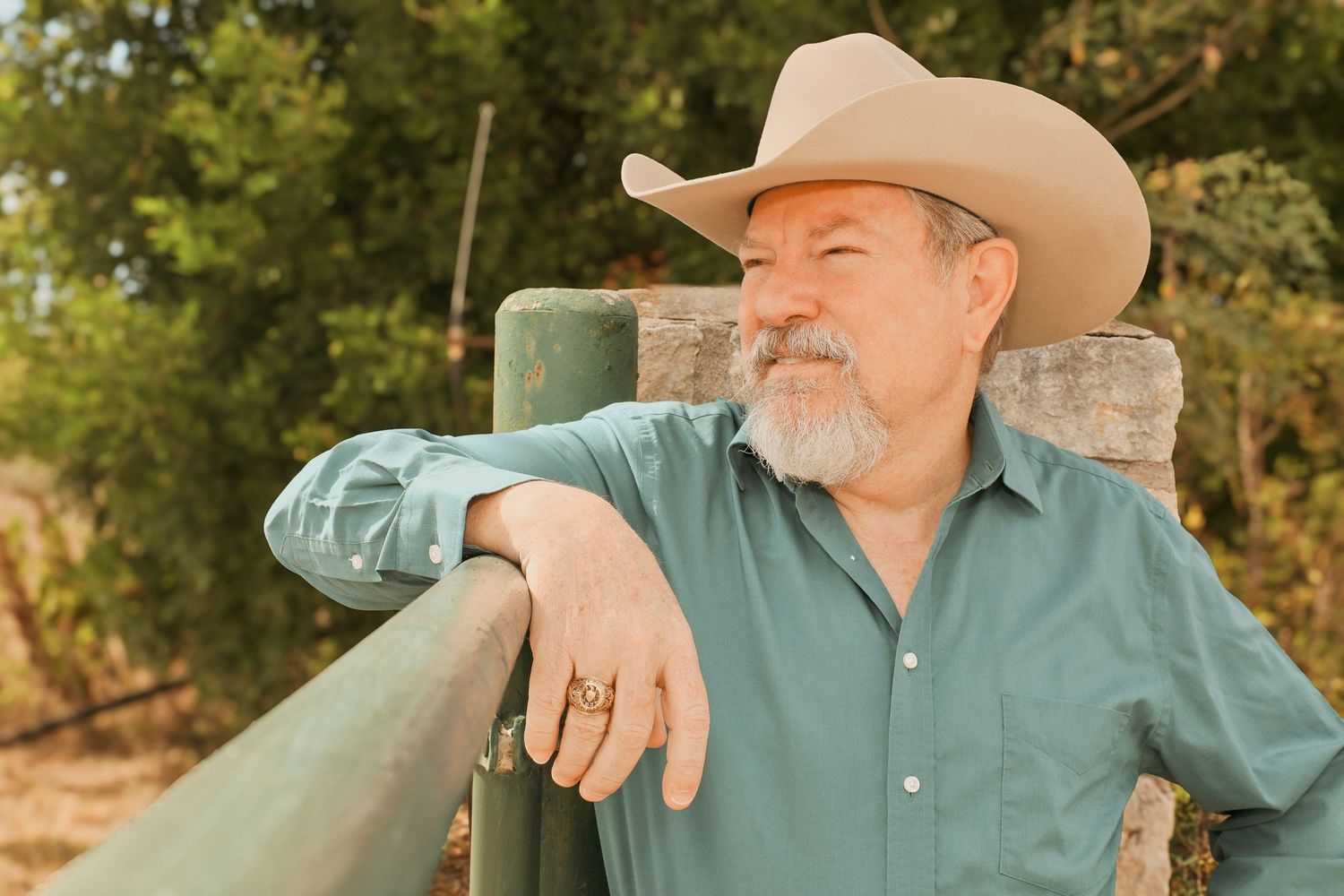 Older man in cowboy hat and teal shirt leaning on green fence in outdoor ranch setting.