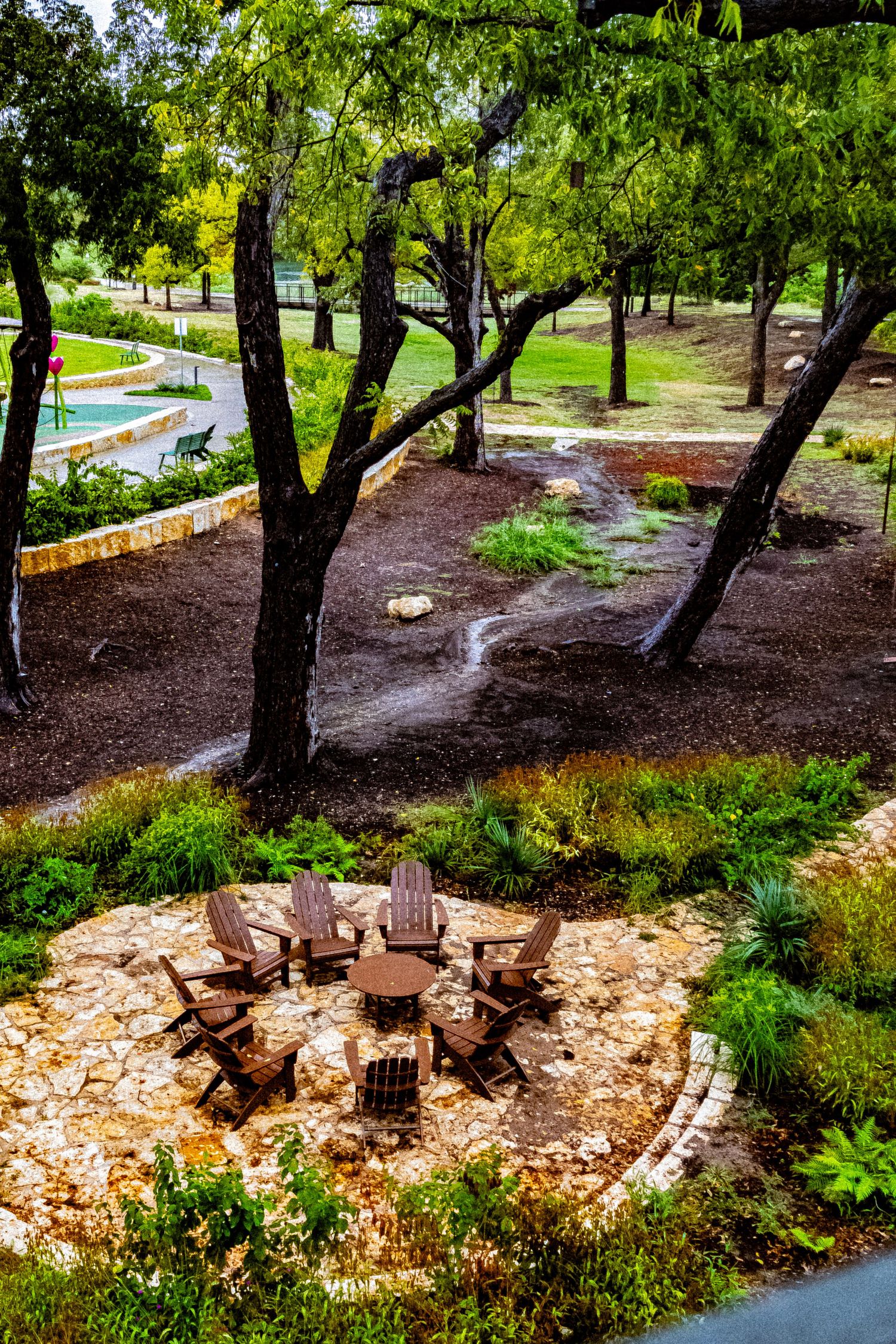 Outdoor stone patio with Adirondack chairs surrounding a fire pit, nestled among trees in a lush park setting.