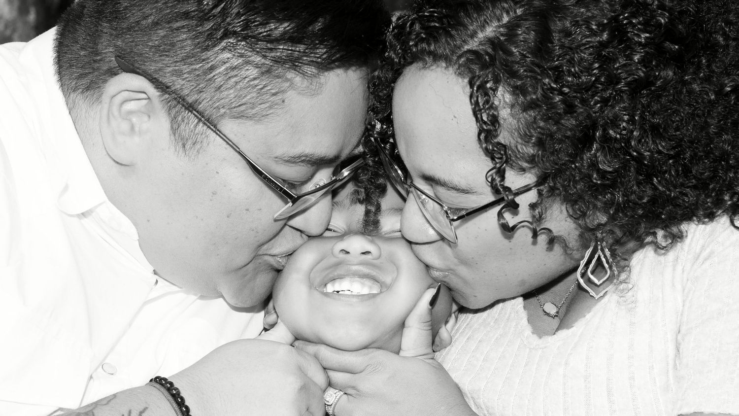 Black and white photo of two parents lovingly kissing their smiling baby on both cheeks simultaneously.