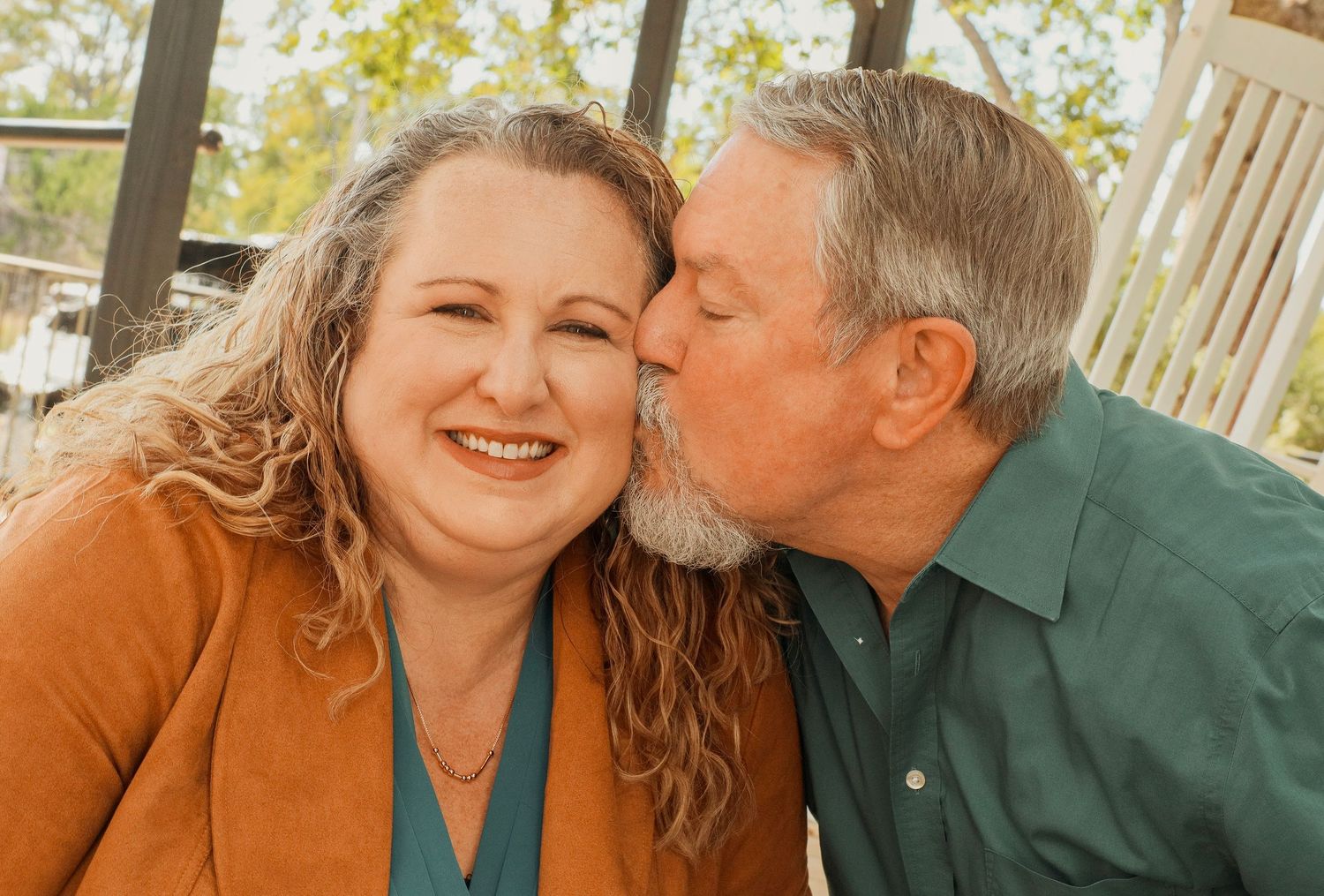 A smiling woman receives a kiss on the cheek from an older bearded man in a teal shirt outdoors.