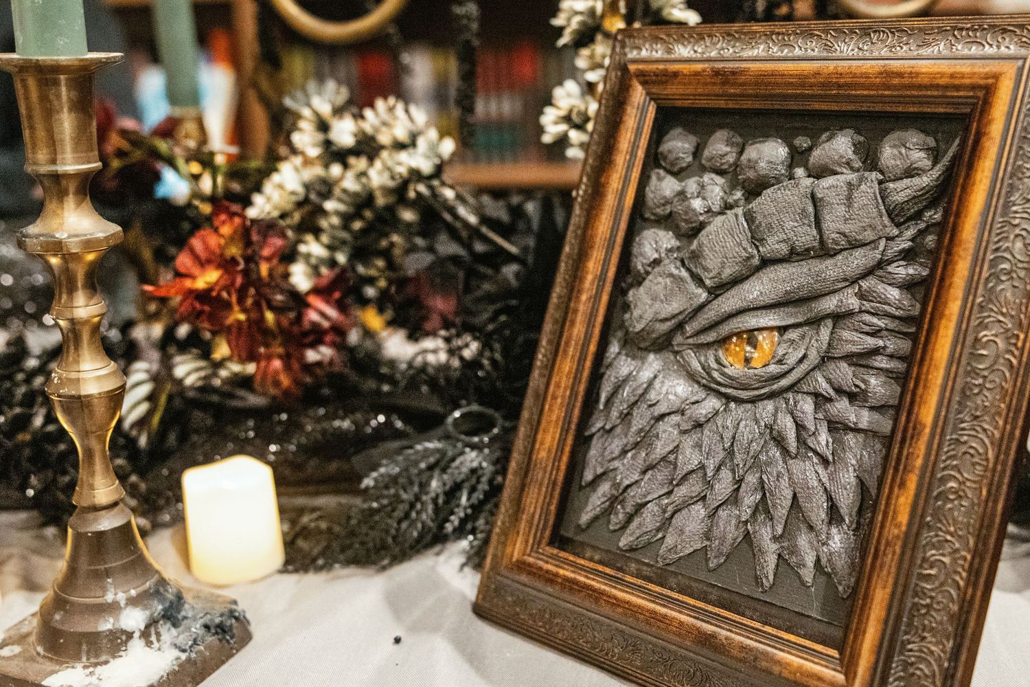 A framed gothic eye artwork with textured dark relief sits beside a brass candlestick and white candle on a decorated table.