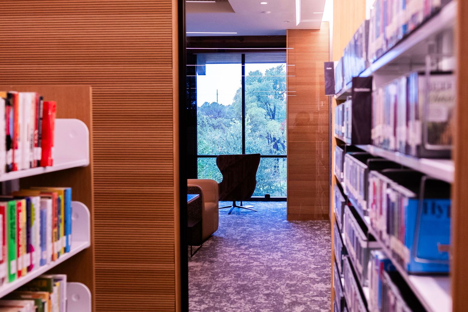 Library aisle with wooden shelves full of books leading to a cozy reading nook with a chair by a large window.