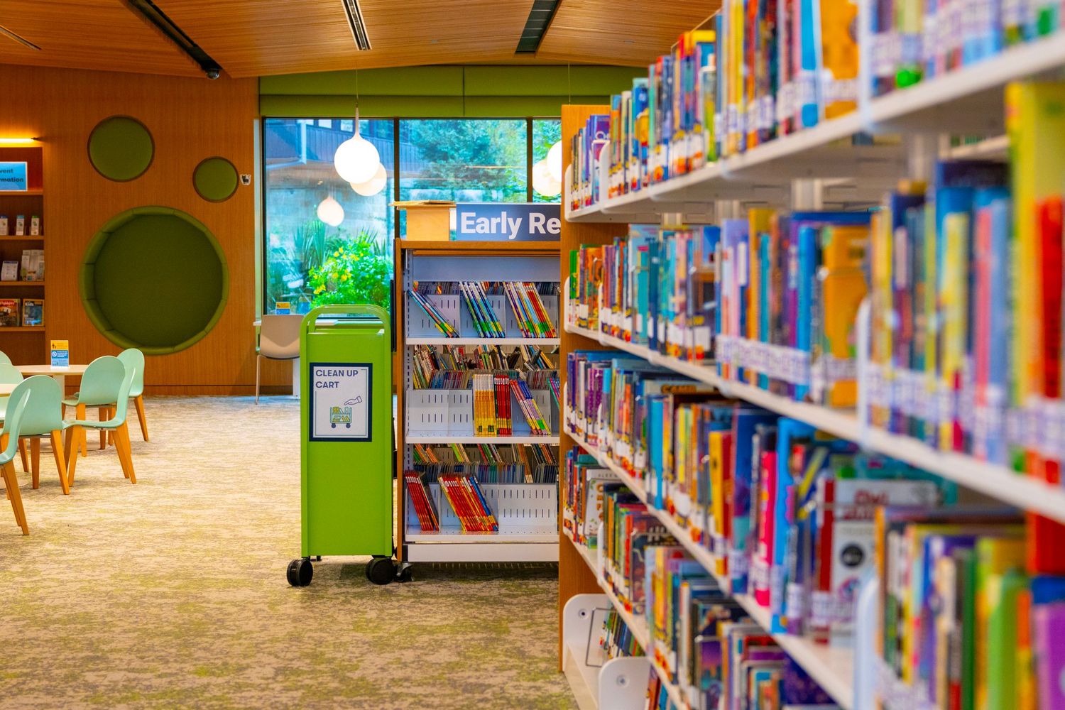 Colorful children's library with shelves of books, green display kiosk, and bright reading area with natural light.