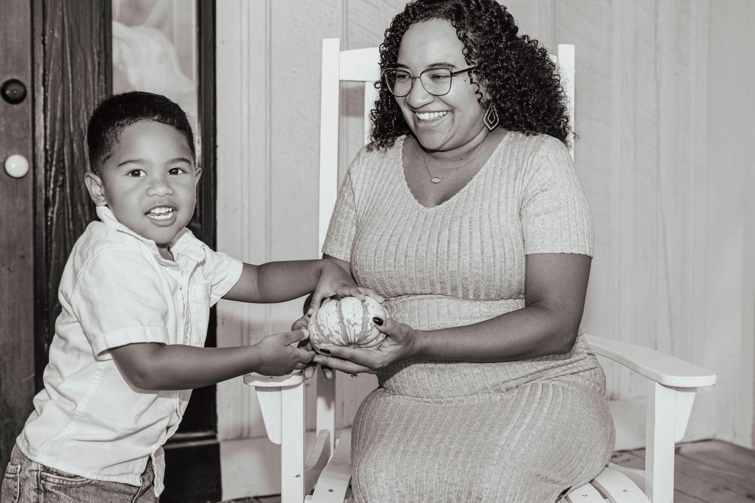 Black and white photo of a smiling woman in glasses and a young boy sharing a pumpkin while seated together indoors.