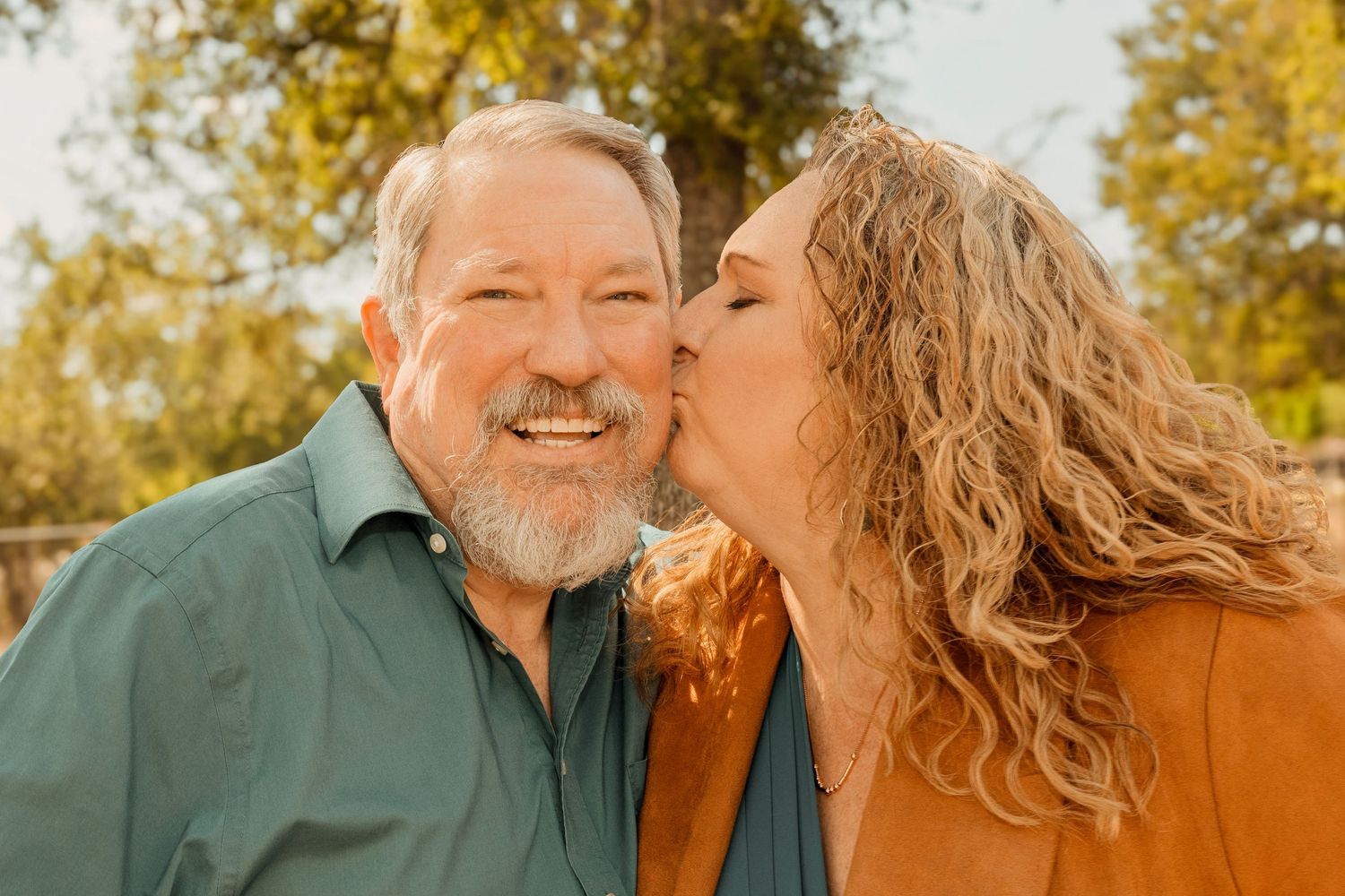 Woman with curly hair kissing smiling bearded man on cheek outdoors with autumn trees in background.