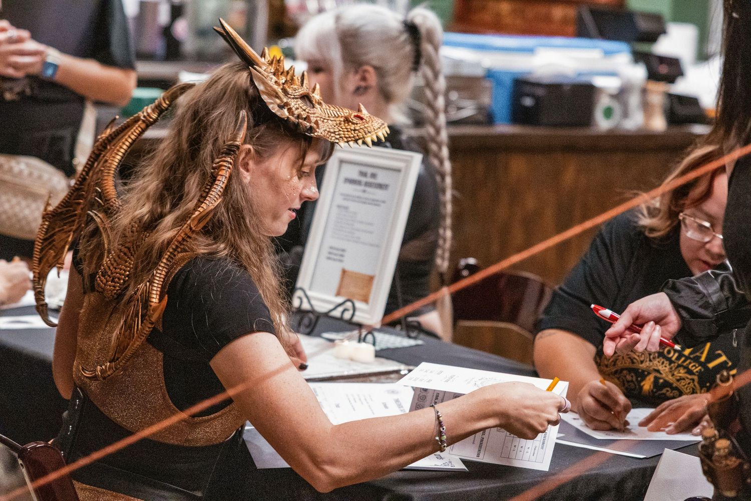 Woman wearing a dragon headpiece signs paperwork at a fantasy convention vendor table.