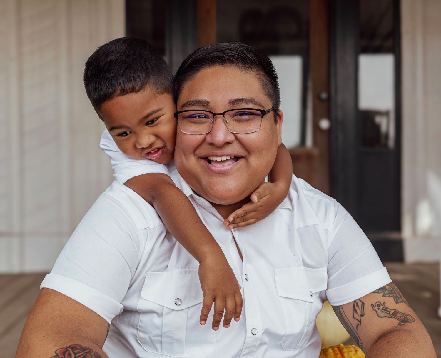 A smiling woman with short hair and glasses hugs a young boy on a porch, both wearing white shirts.