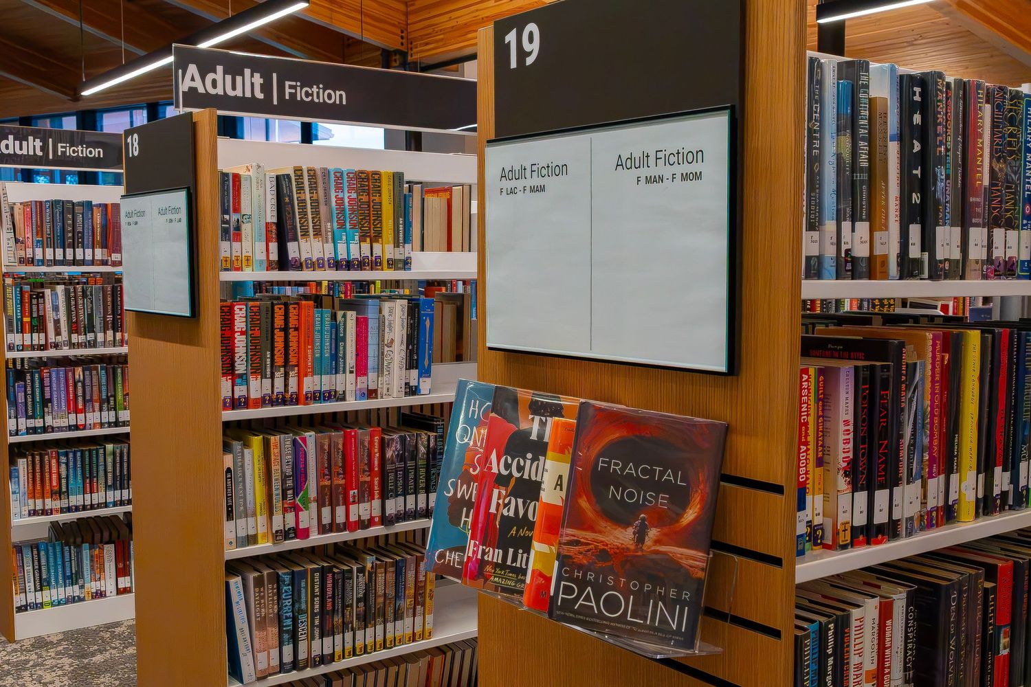 Adult fiction section of a library with colorful books on shelves, including Fractal Noise by Christopher Paolini.