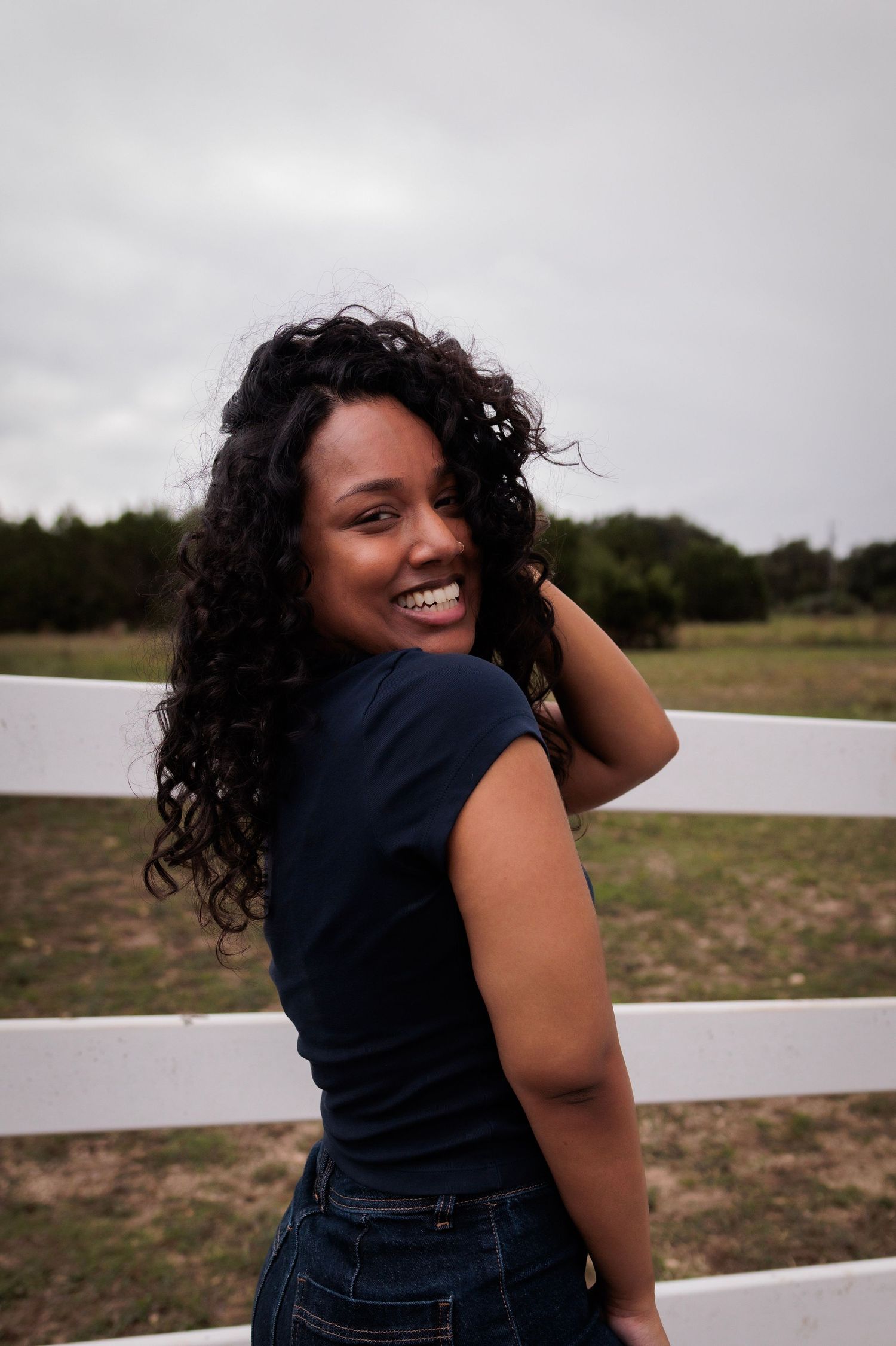 Smiling young woman with curly hair in navy shirt and jeans leaning against white fence in rural field.