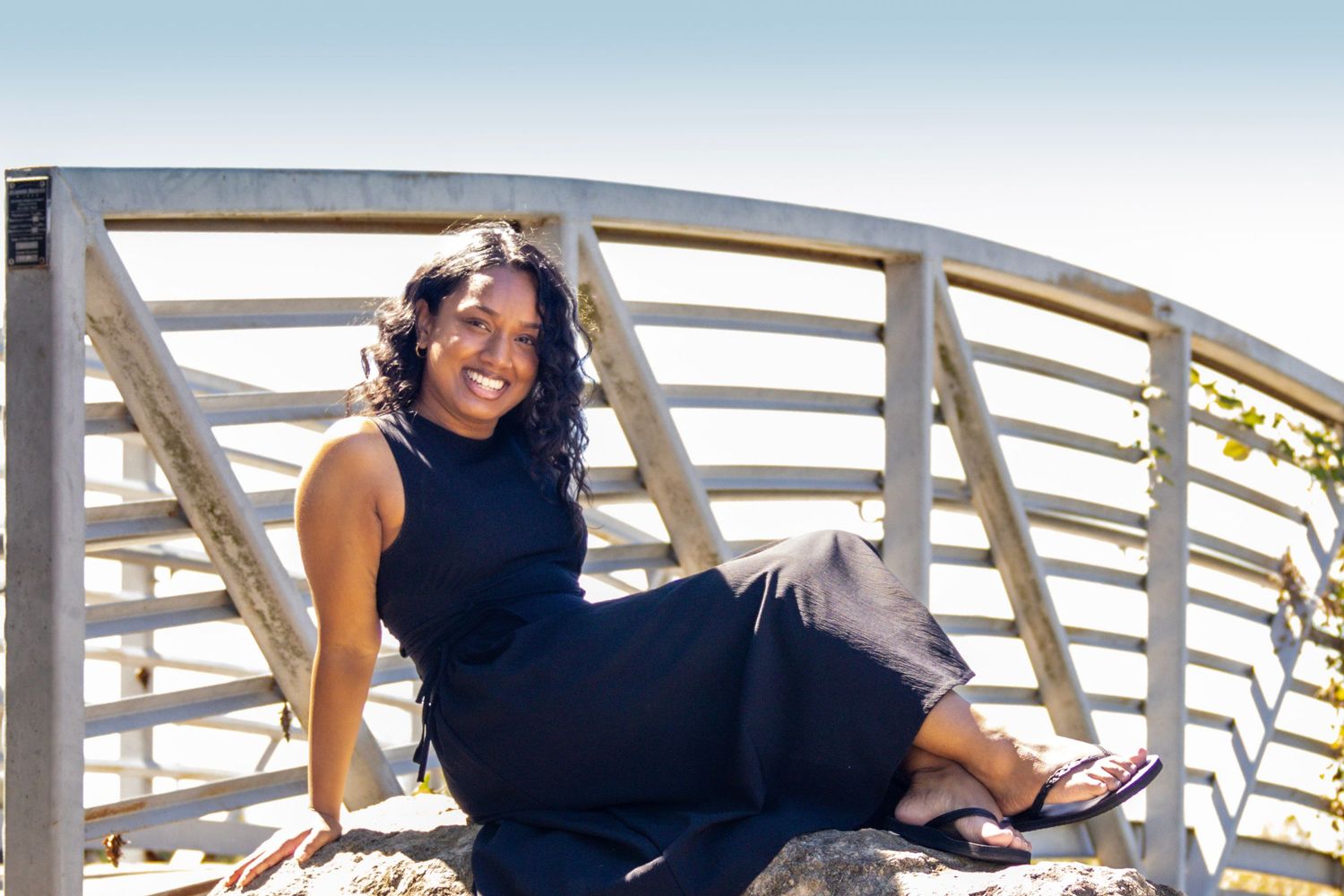 Smiling woman in black dress sitting on rocks near decorative metal railing outdoors on a sunny day.