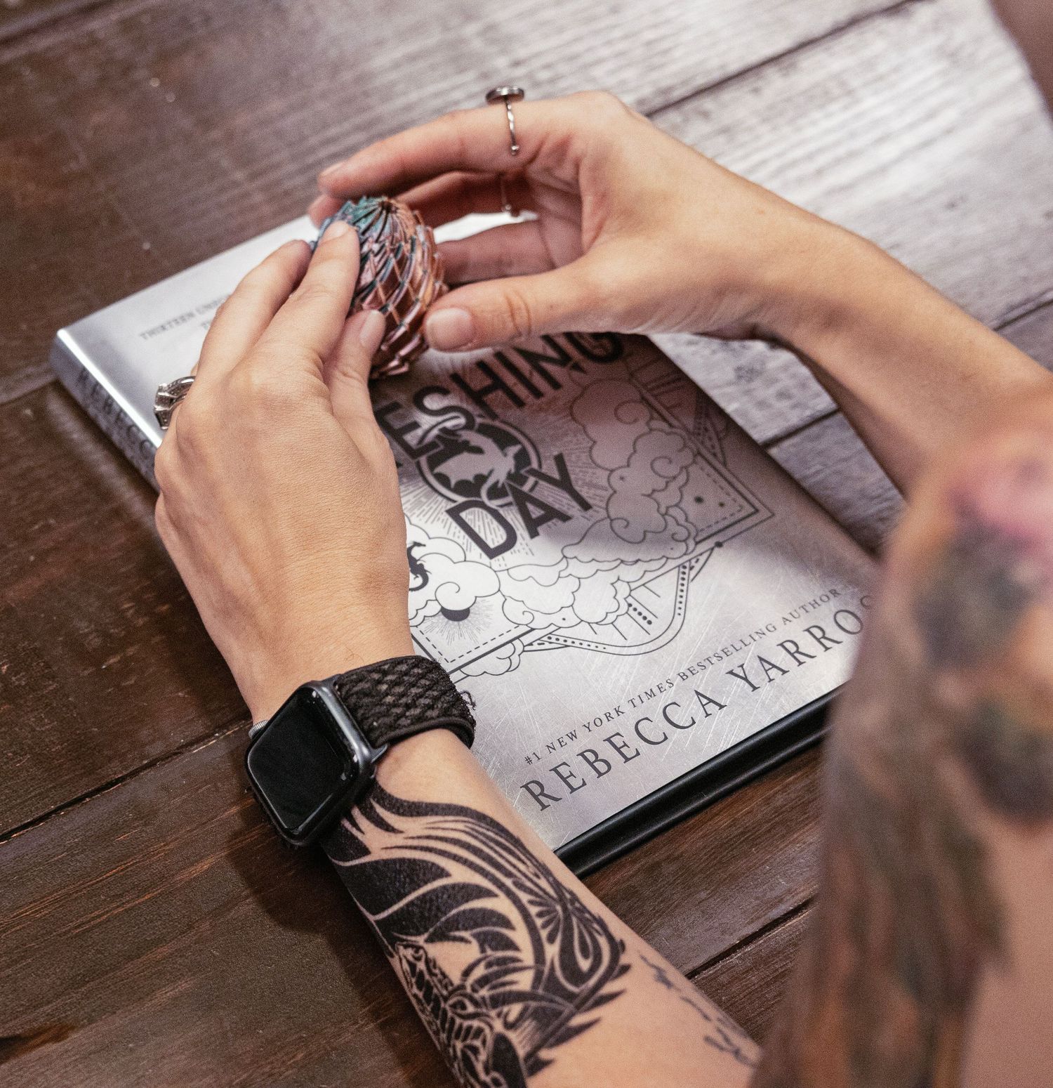 Tattooed hands holding a bookmark over Rebecca Yarros book on wooden table, person wearing Apple Watch.