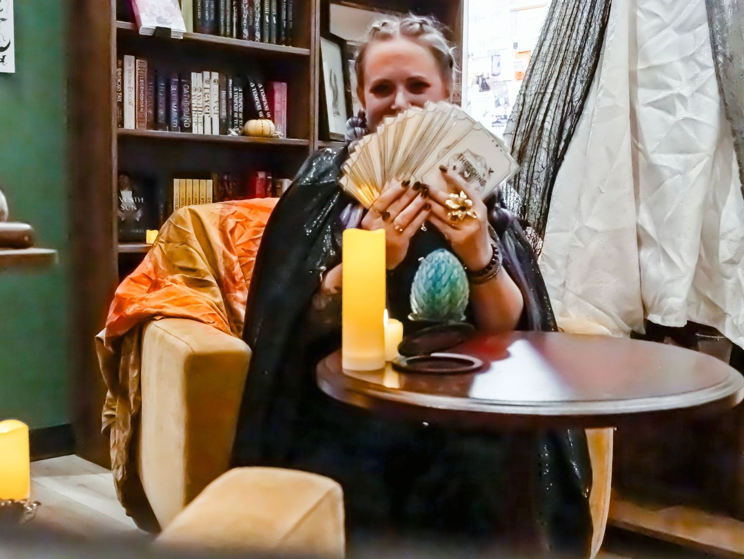 A fortune teller fans tarot cards at a candlelit table with mystical decor and bookshelves in the background.
