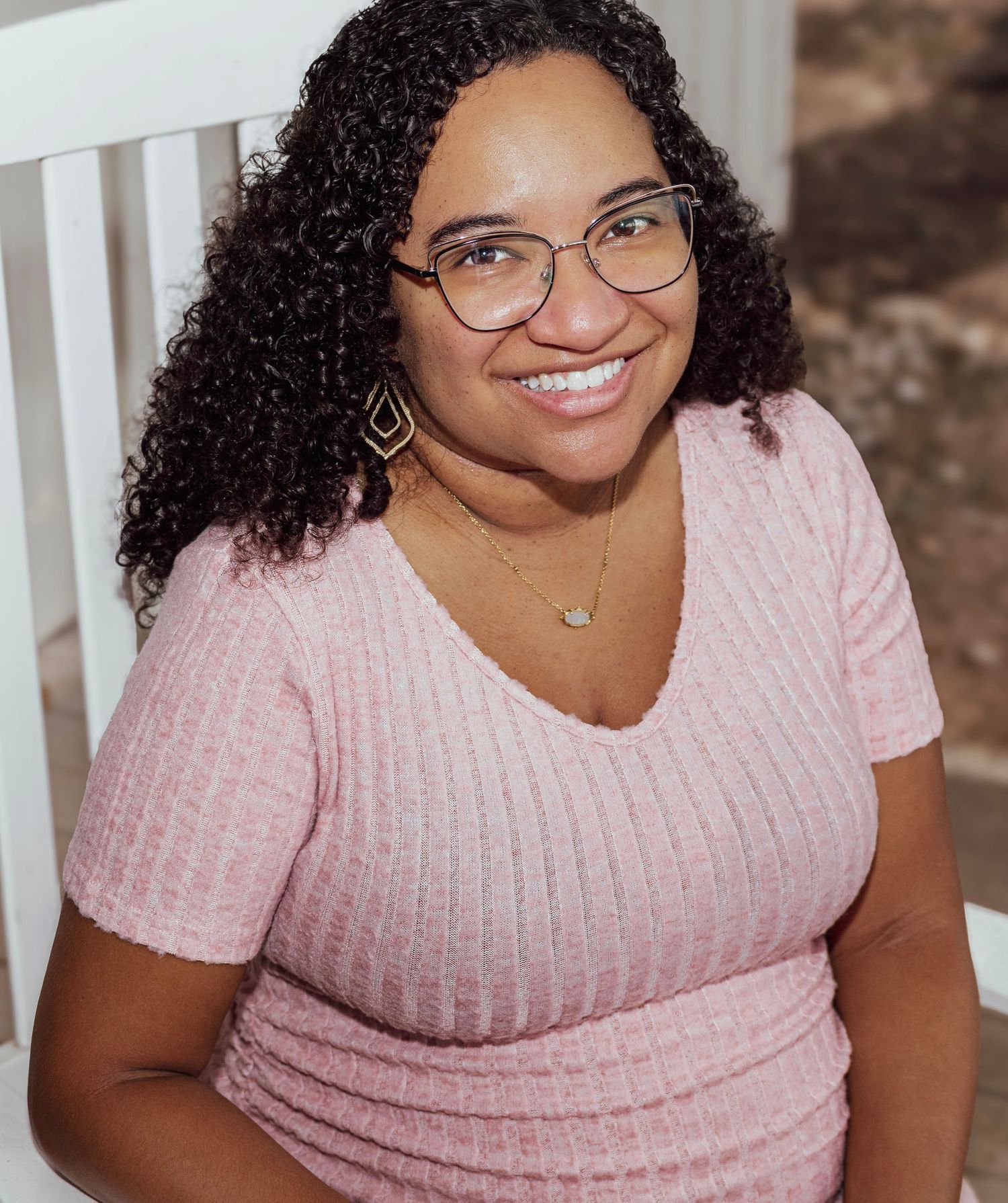 Smiling woman with curly hair and glasses wearing a pink ribbed top, seated outdoors in a warm portrait photo.