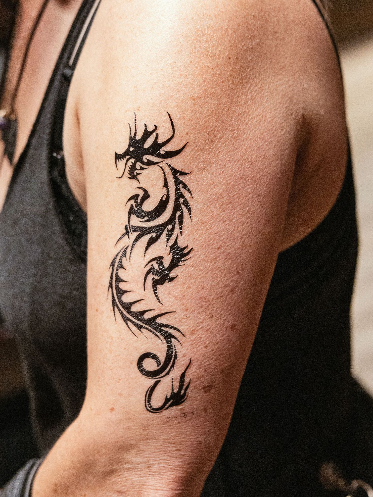 Close-up of a tribal black dragon tattoo on a person's upper arm wearing a black tank top.