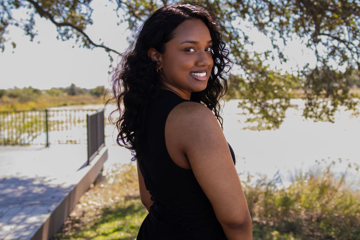 A young Black woman in a black dress smiles over her shoulder outdoors near a scenic waterway.